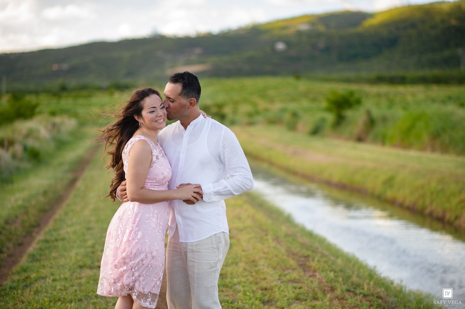 Couple embracing and smiling in a lush green field with a stream and mountains in the background.