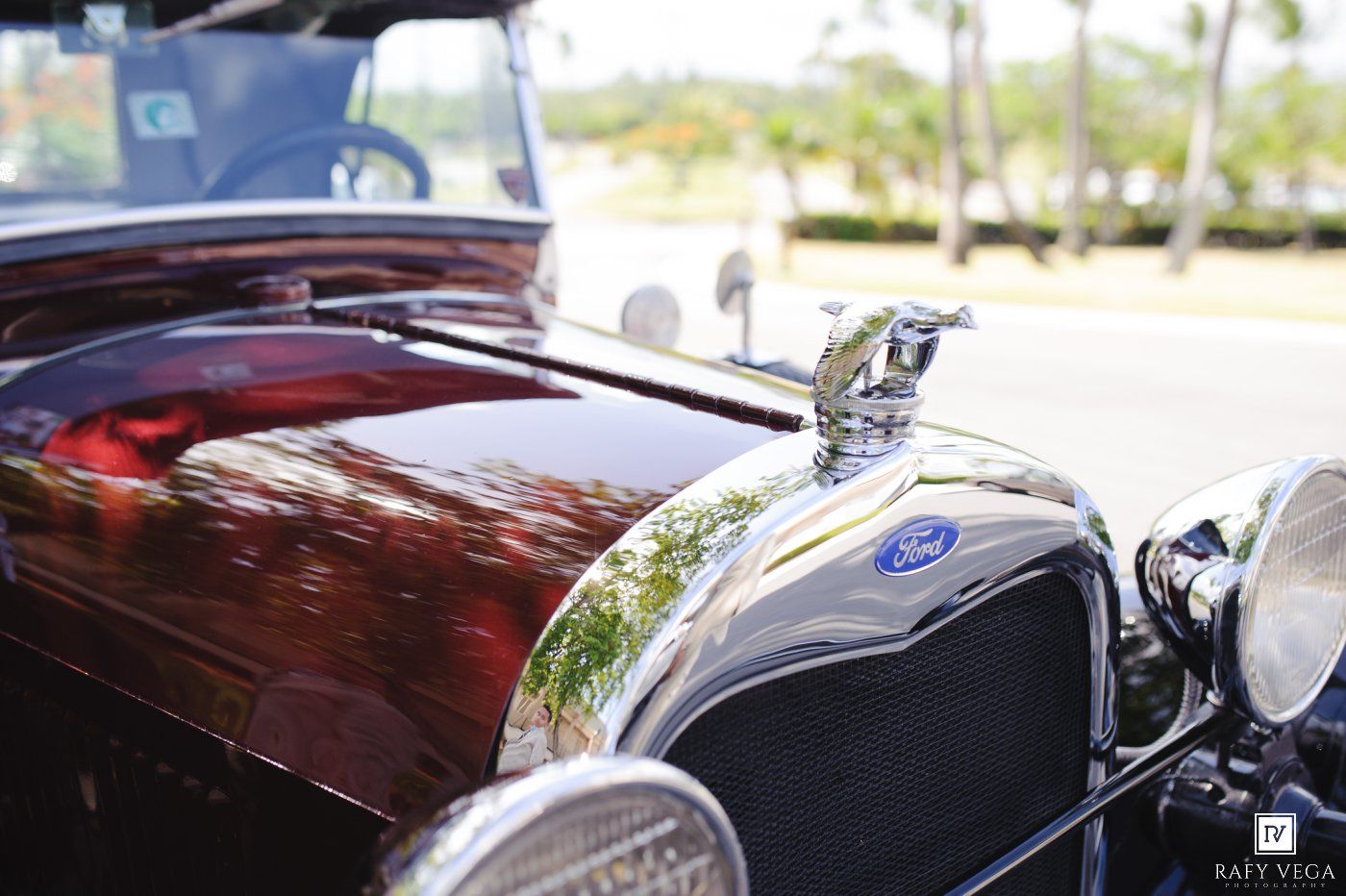 Close-up of a vintage Ford car hood ornament on a shiny dark red classic automobile at an outdoor show.