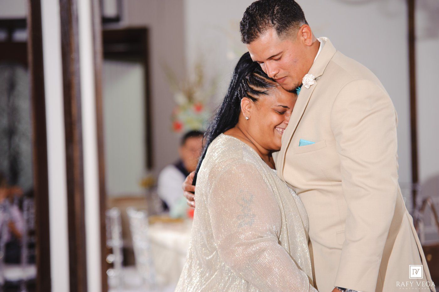 A couple shares a tender embrace during their wedding reception, the bride in white lace and groom in a beige suit.