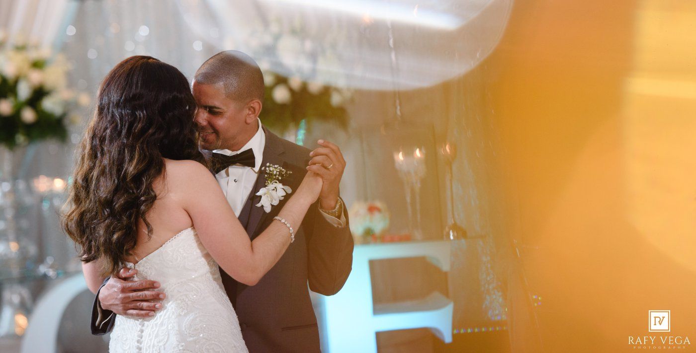 A bride and groom share their first dance, embracing romantically in a warmly lit wedding reception venue.