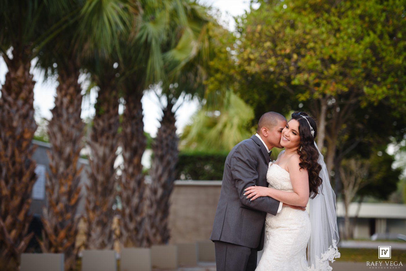 Bride and groom share a romantic kiss outdoors surrounded by palm trees on their wedding day.