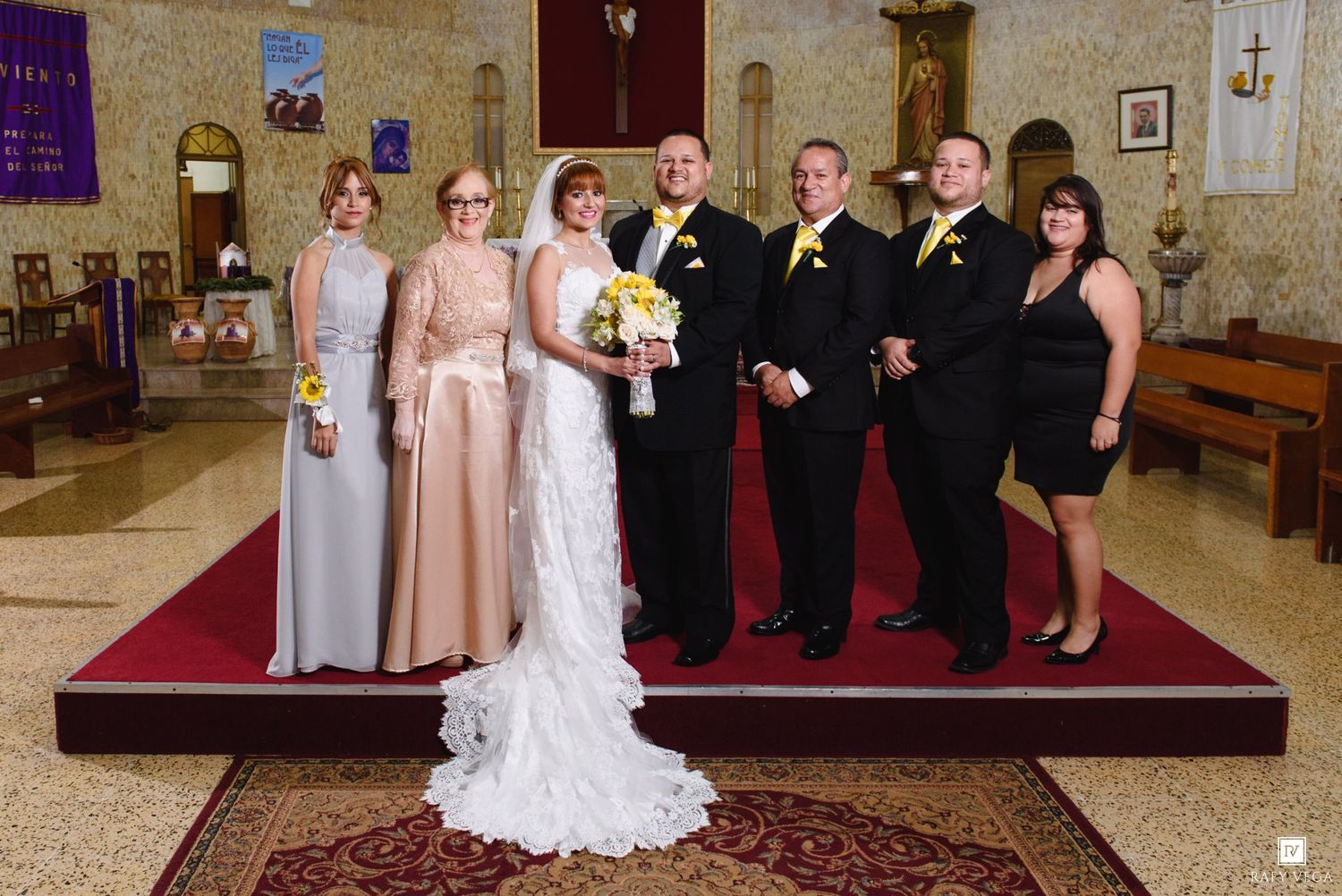 Wedding party posing on church altar steps with bride holding yellow bouquet, bridesmaids and groomsmen in formal attire.