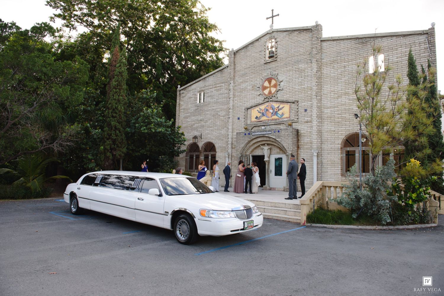 White stretch limousine parked outside a stone church during a wedding celebration with guests gathered at the entrance.