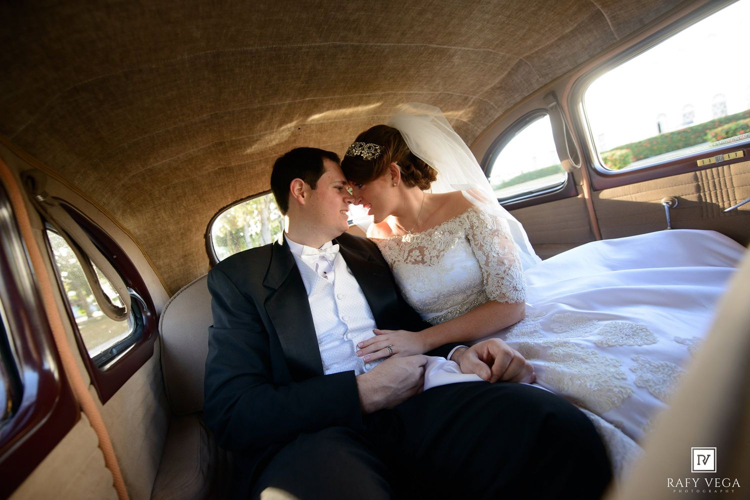Bride and groom share a romantic moment inside a vintage wedding car, dressed in elegant bridal gown and tuxedo.