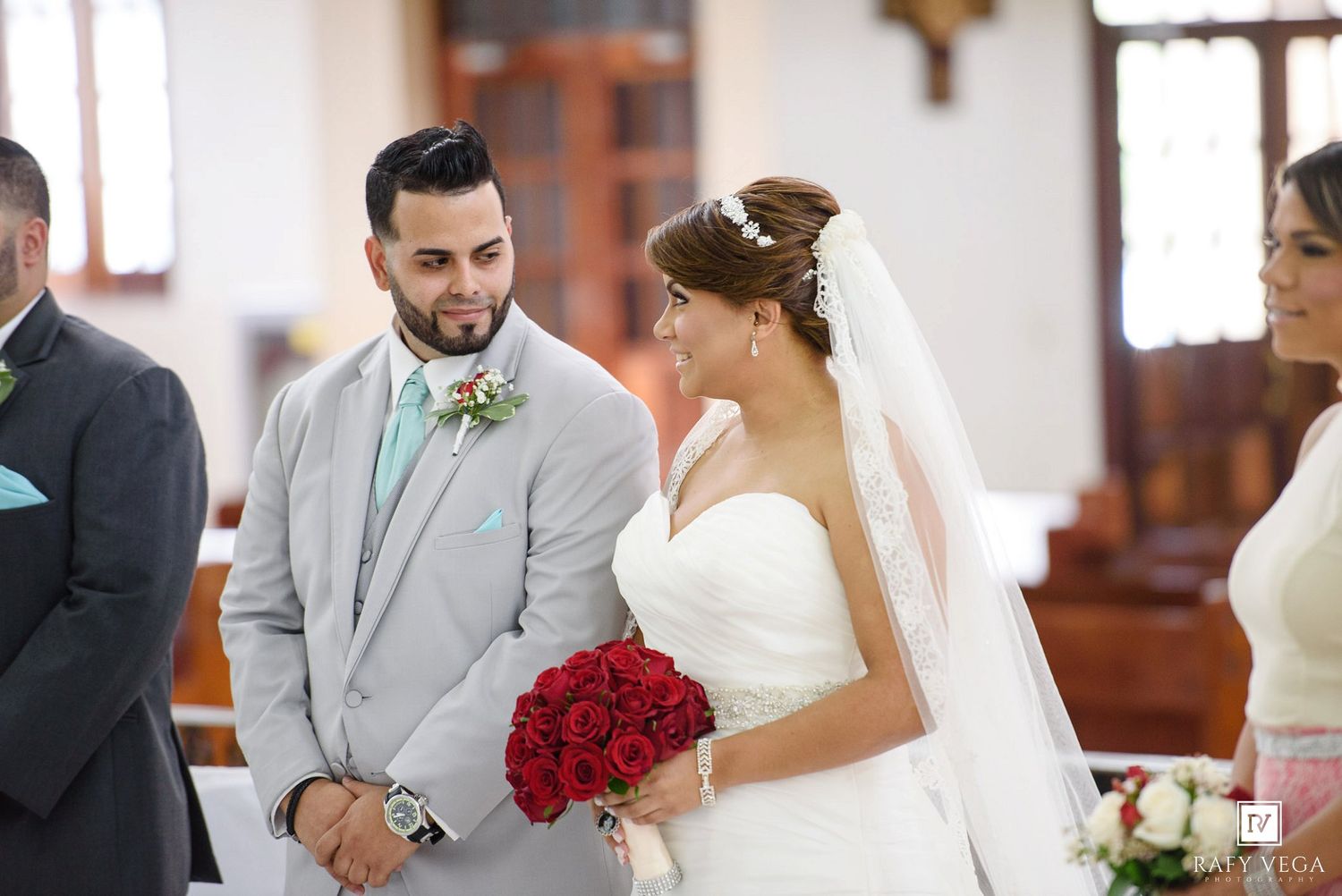 Bride holding red roses bouquet stands beside groom in gray suit during wedding ceremony inside a church.