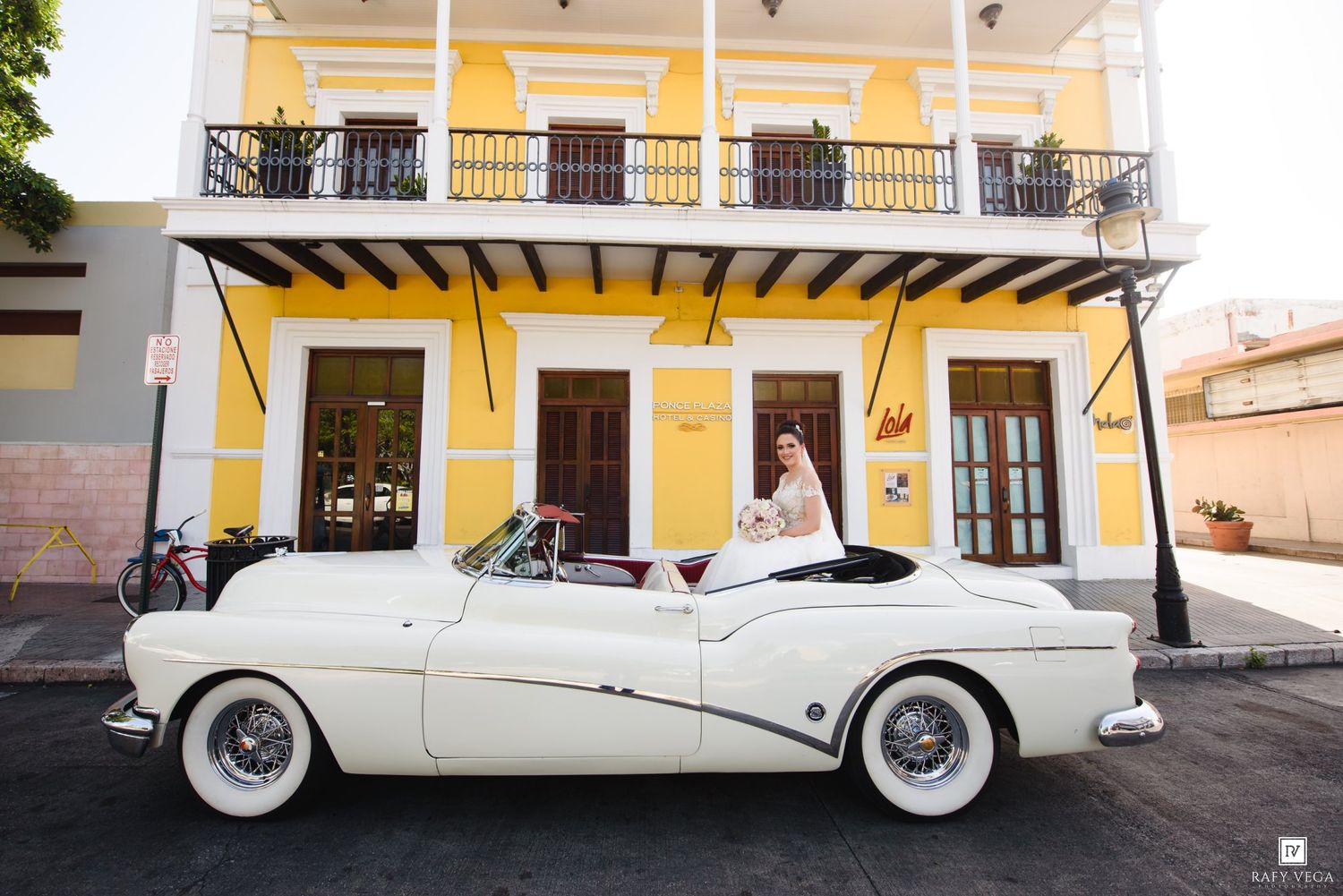 Bride posing in a white vintage convertible car in front of a yellow colonial building in Old San Juan.