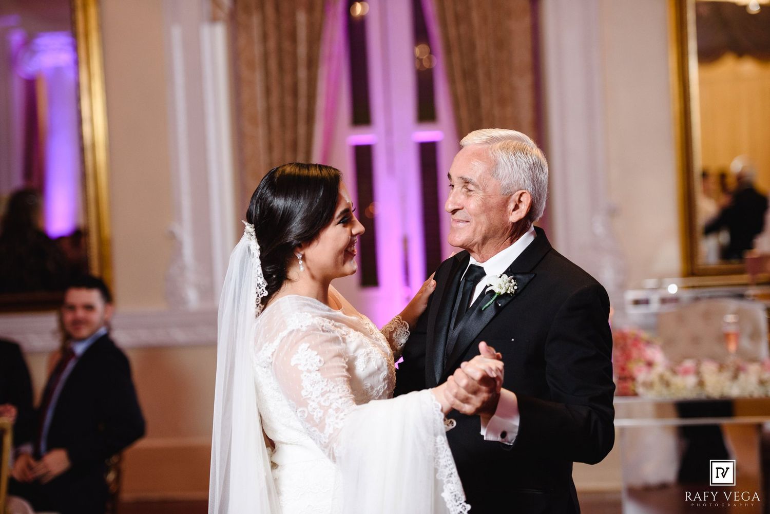 Bride in white lace gown shares father-daughter dance at elegant wedding reception with pink floral decor.