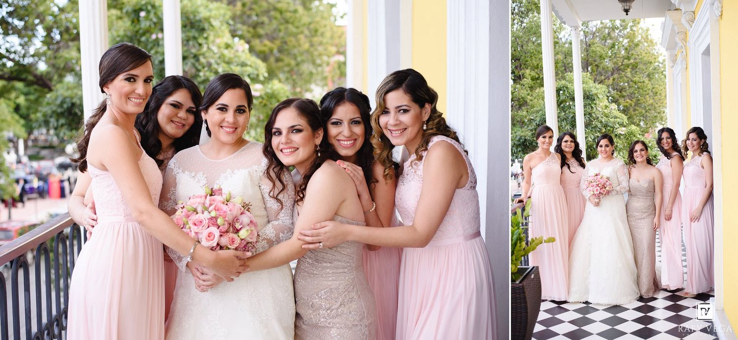 Bride poses with bridesmaids in pink gowns on a decorated porch, holding a bouquet of pink and white flowers.