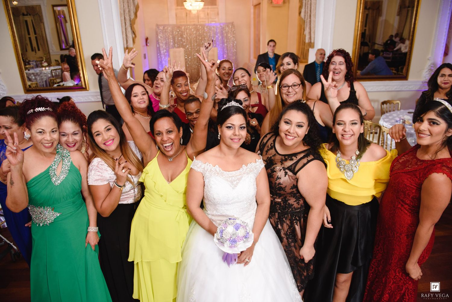 Bride in white gown poses with smiling bridesmaids and guests in colorful dresses at a wedding reception.