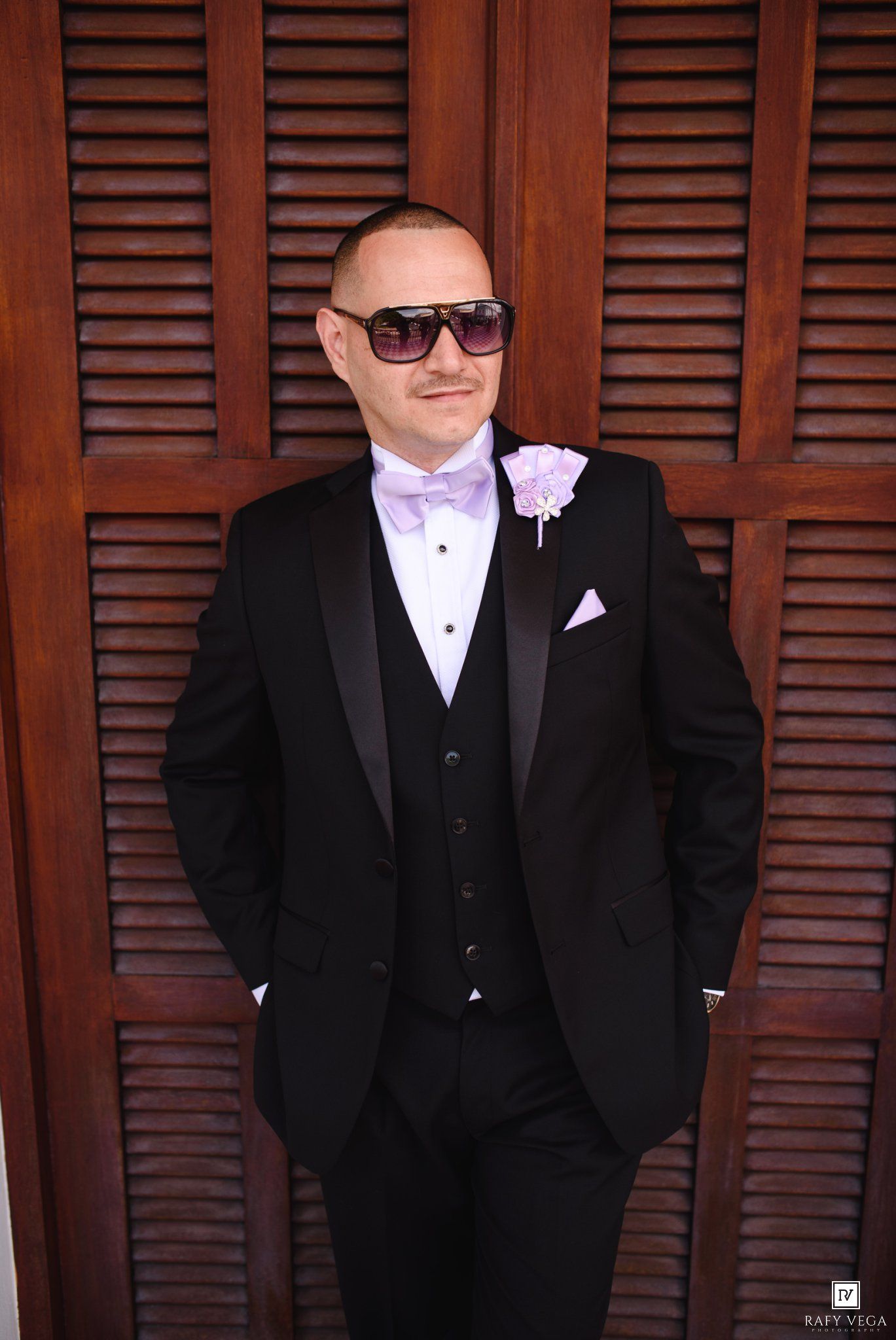 Groom in black tuxedo with lavender bow tie, boutonniere, sunglasses, posing against wooden shutters.