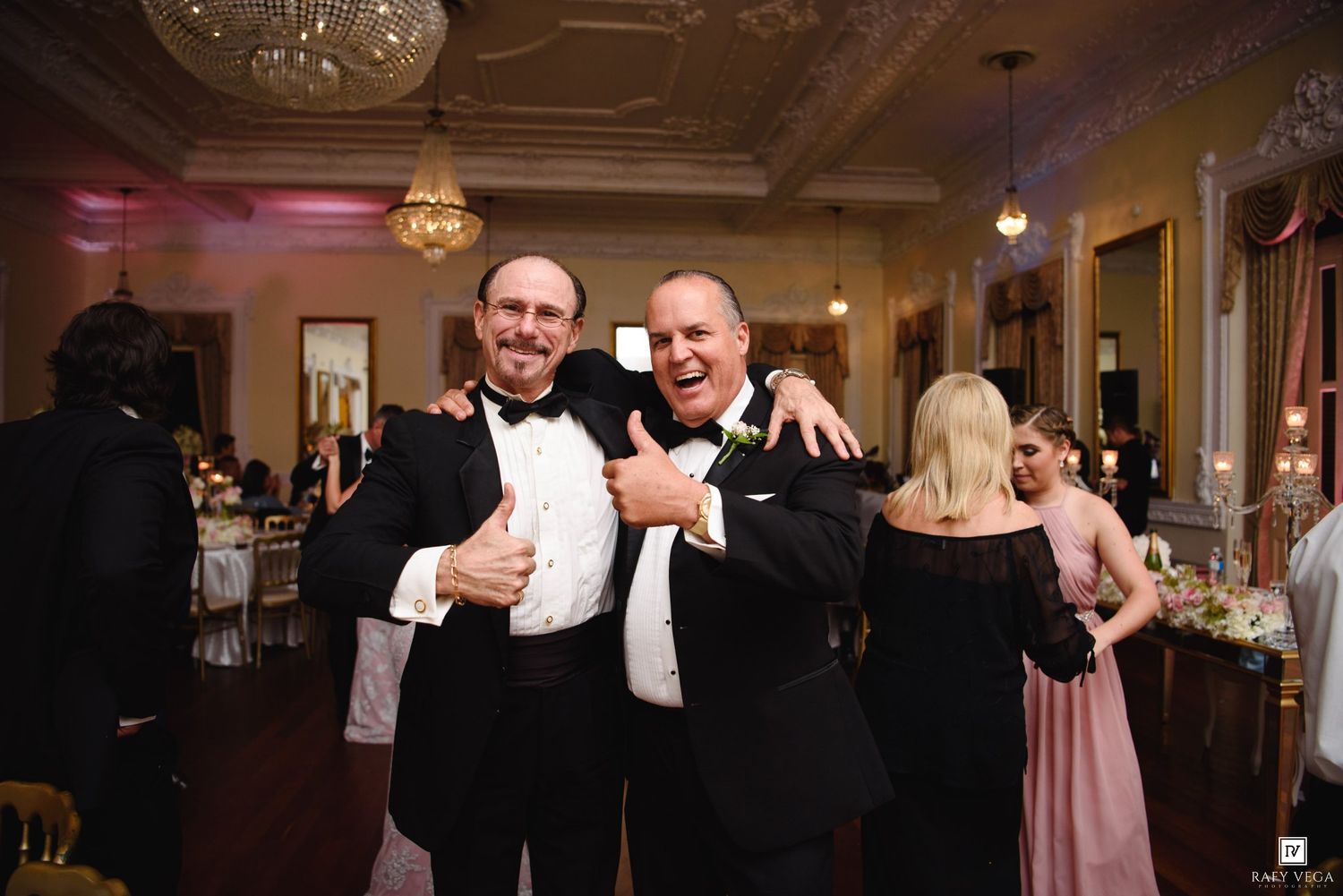 Two men in tuxedos smile and pose together at a formal wedding reception in an elegant ballroom.