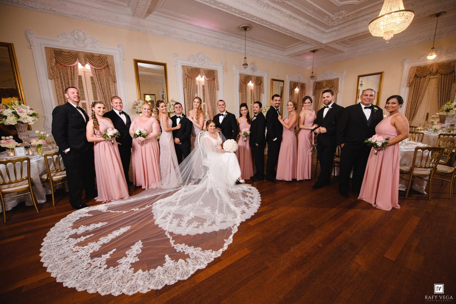 A wedding party poses in an elegant ballroom, bridesmaids in pink gowns flanking the bride in a cathedral veil.
