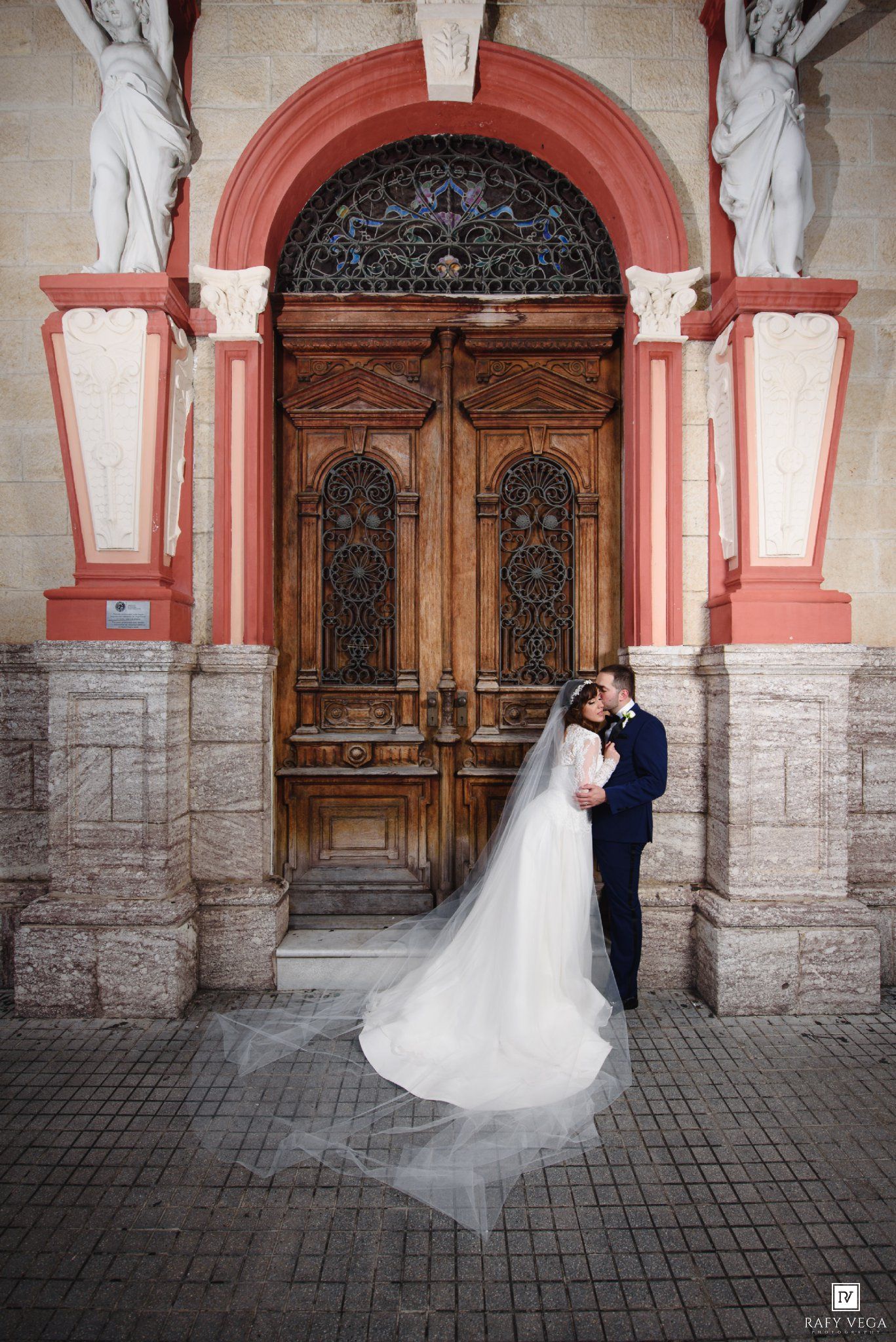Bride and groom kissing before ornate wooden doors of a pink stone church, with flowing white veil.