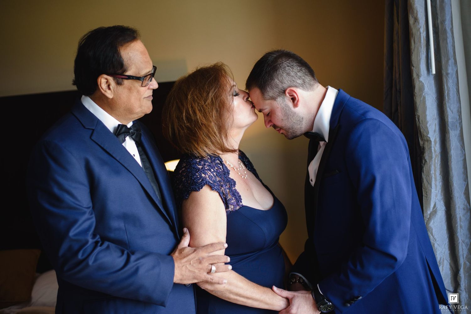 Groom in blue suit shares tender moment with parents in elegant blue attire before wedding ceremony.