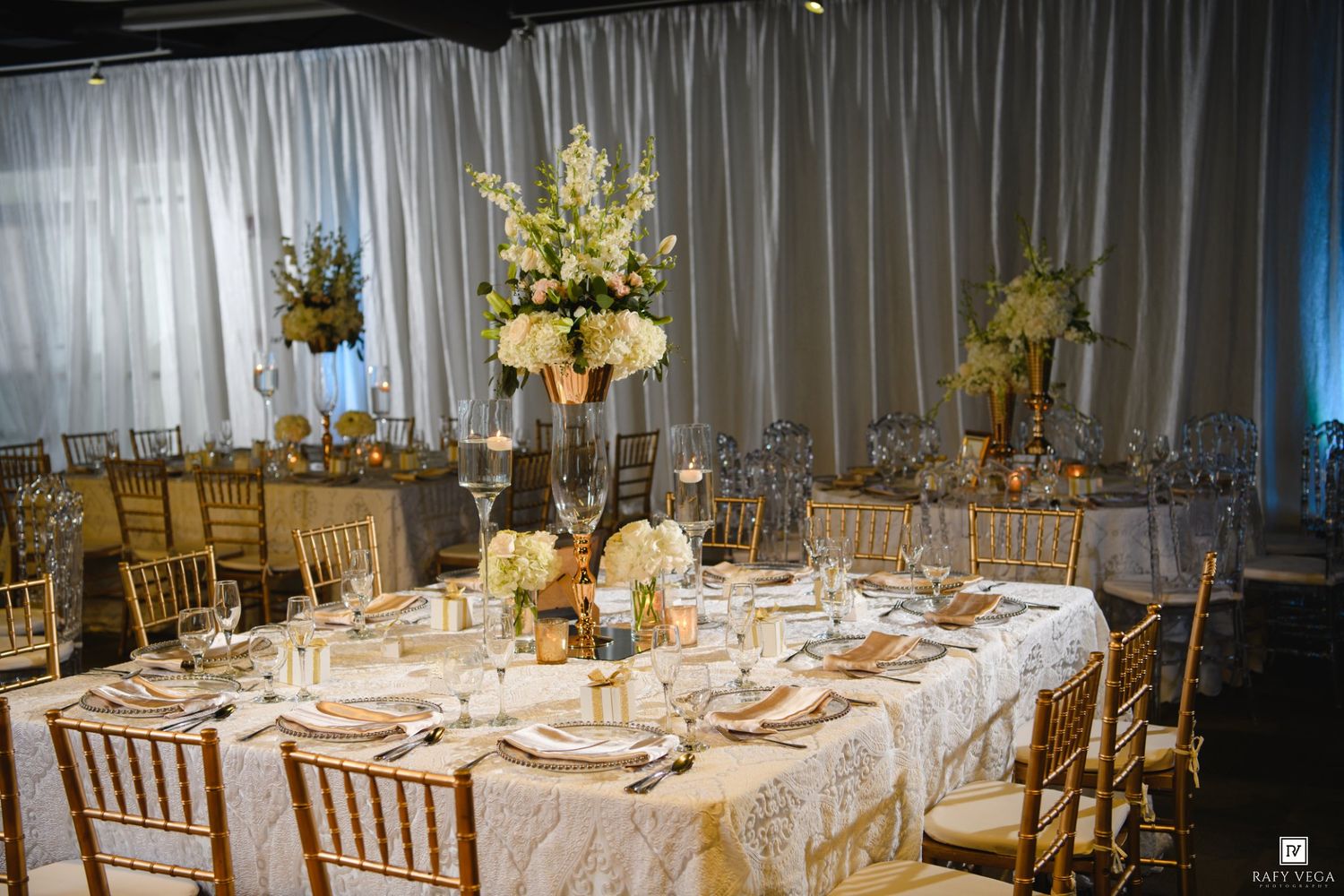 Elegant wedding reception table with gold Chiavari chairs, white floral centerpieces, and white linen tablecloths in a decorated venue.