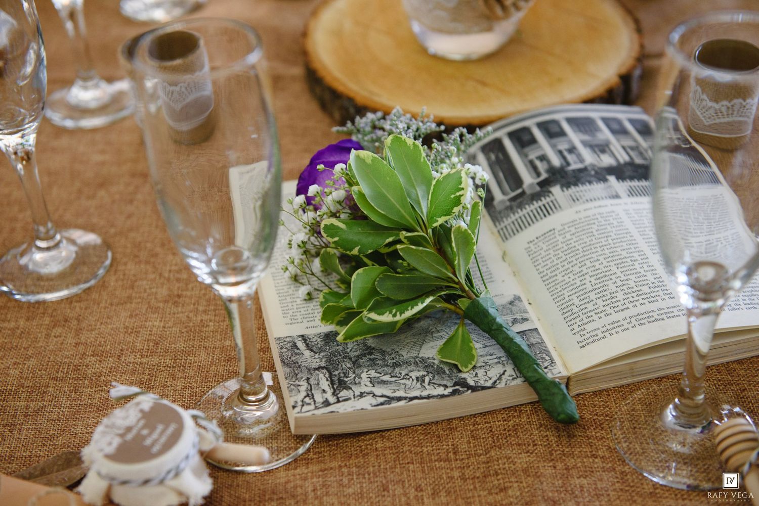 Open book centerpiece with green boutonniere and purple flower, surrounded by champagne flutes on burlap table runner.