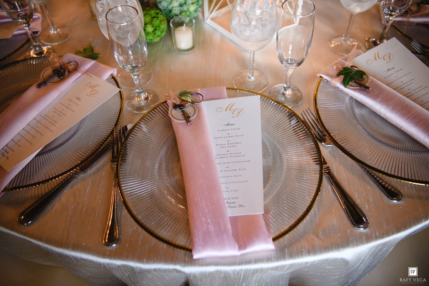 Elegant wedding table setting with pink menu cards, gold charger plates, crystal glasses, and blush napkins on a silver tablecloth.
