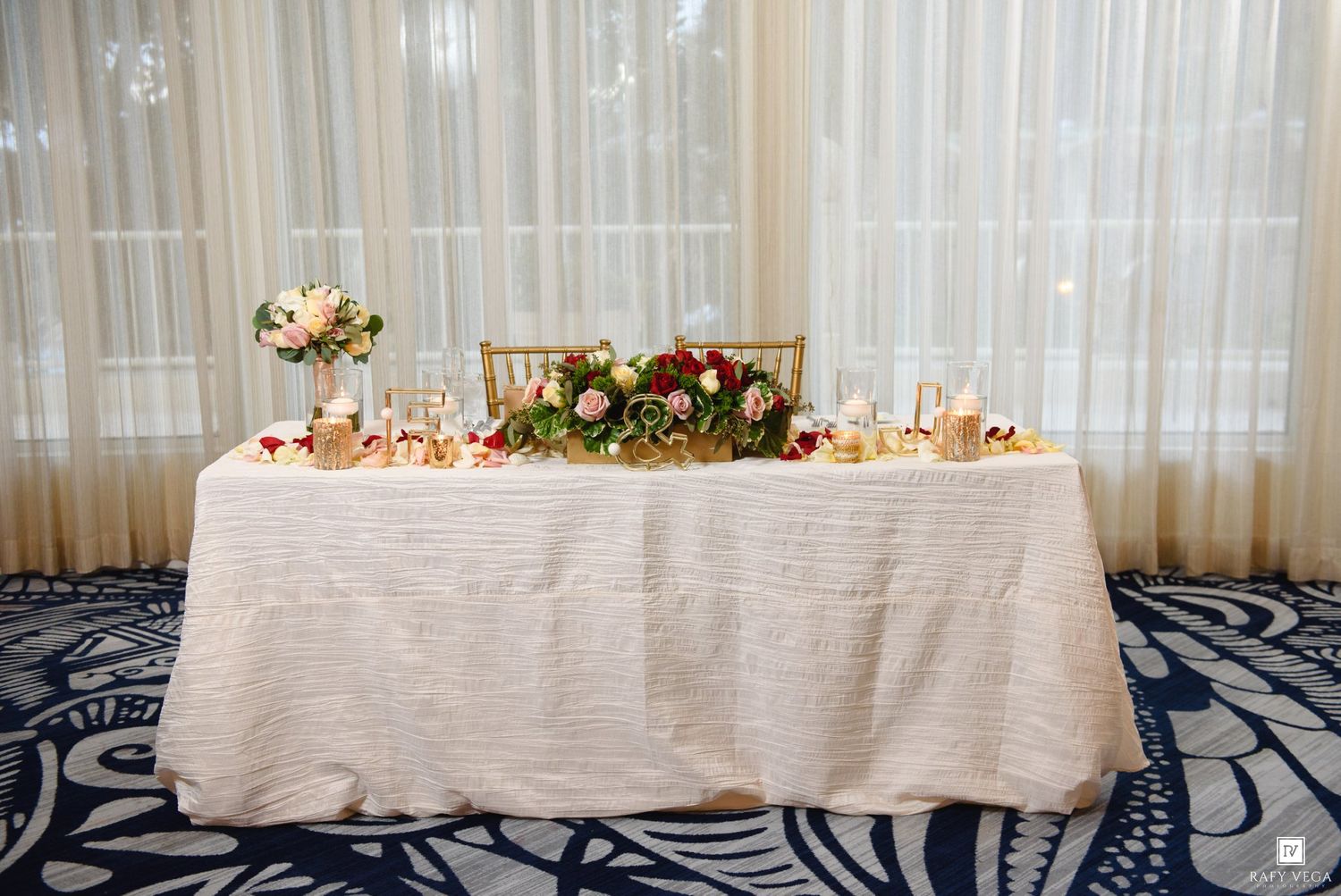 Elegant wedding sweetheart table with white linen, floral garland centerpiece, candles, and sheer curtain backdrop.