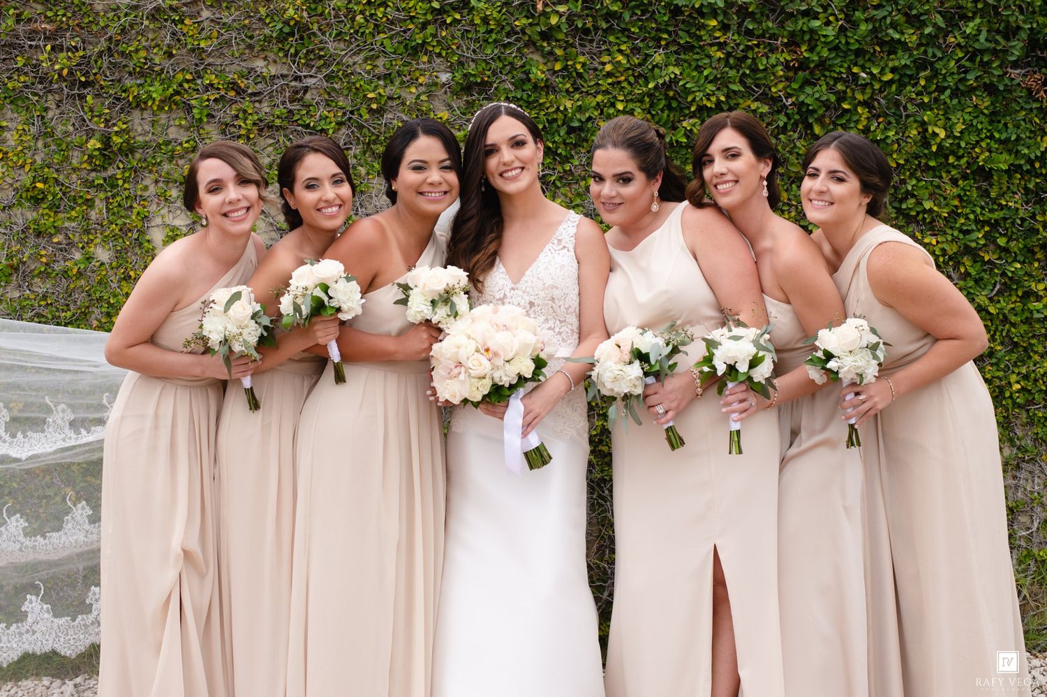 Bride in white gown poses with five bridesmaids in champagne dresses holding white bouquets before ivy wall.