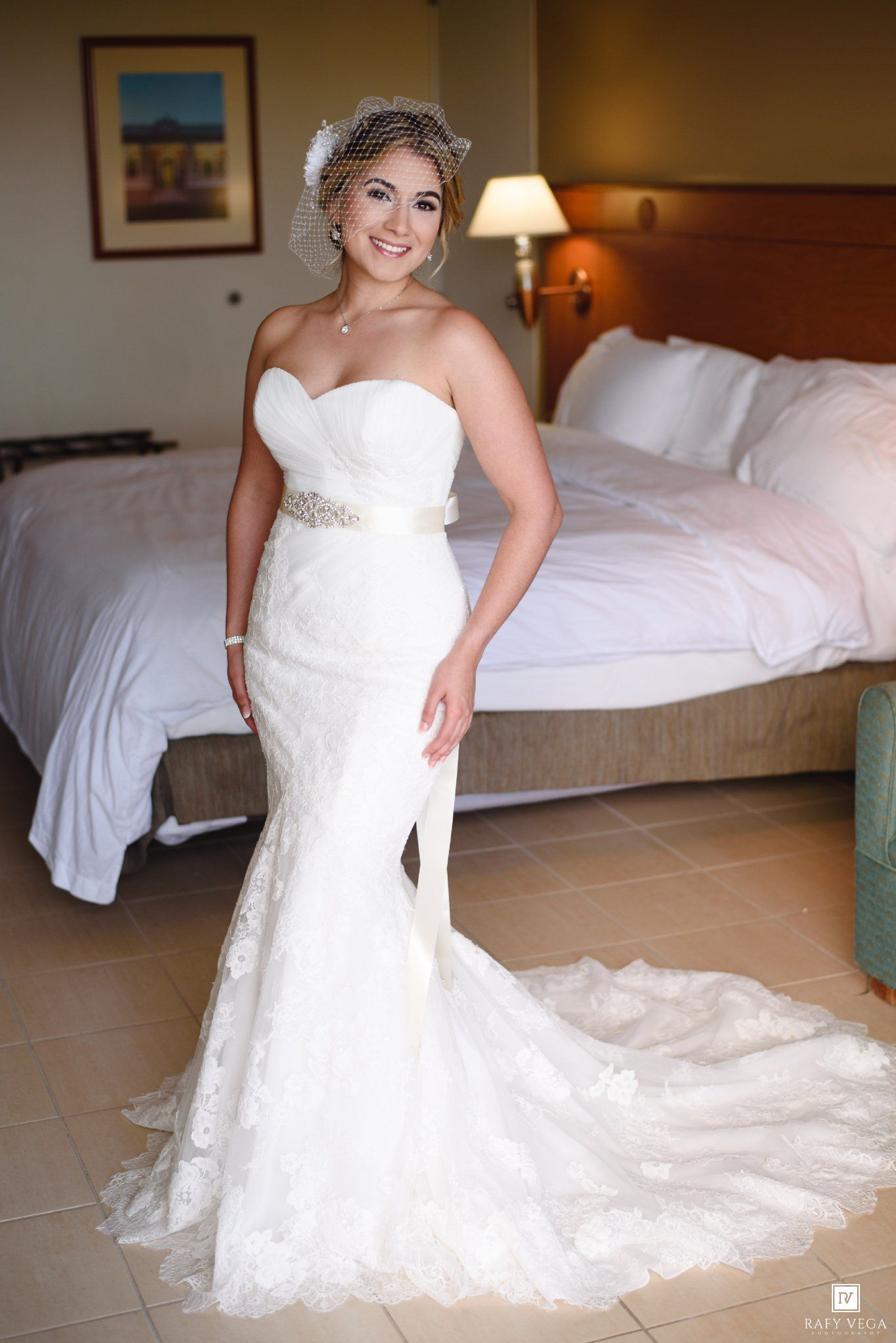 Bride in strapless lace mermaid wedding gown with sash and birdcage veil poses in hotel room.