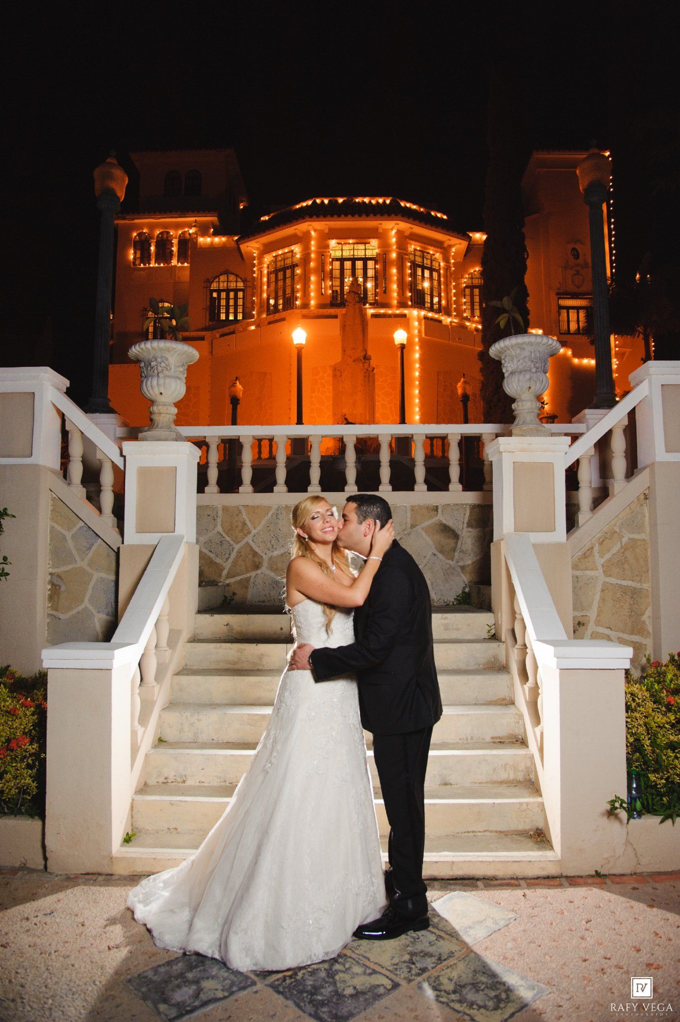 Bride and groom share a romantic kiss on elegant white stairs outside a beautifully lit mansion at night.