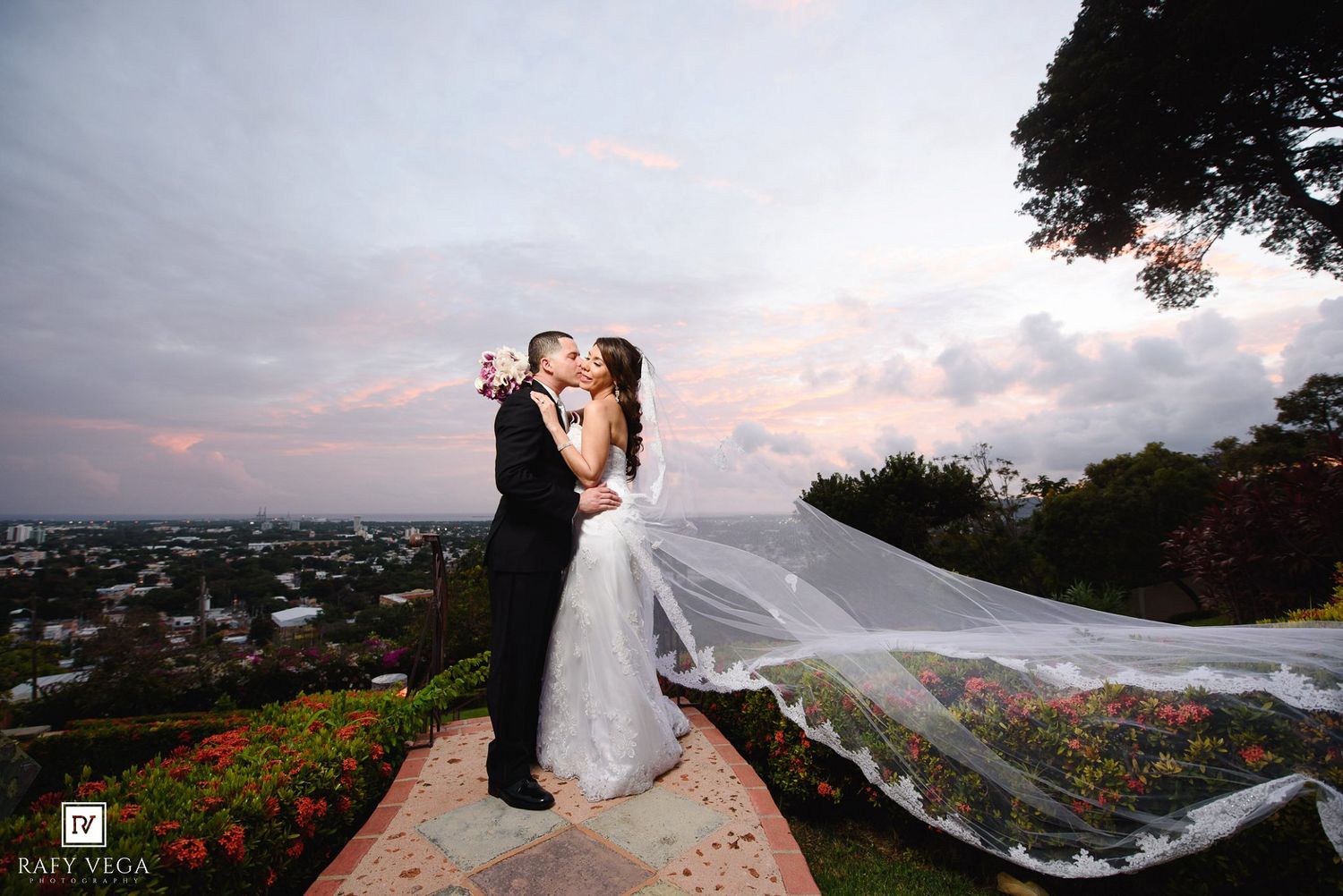 Bride and groom share a romantic kiss at sunset with a flowing veil and city skyline backdrop.