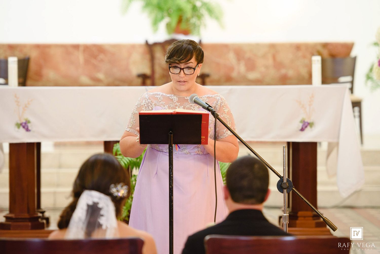 Woman in lavender dress reads at church podium during wedding ceremony at altar.