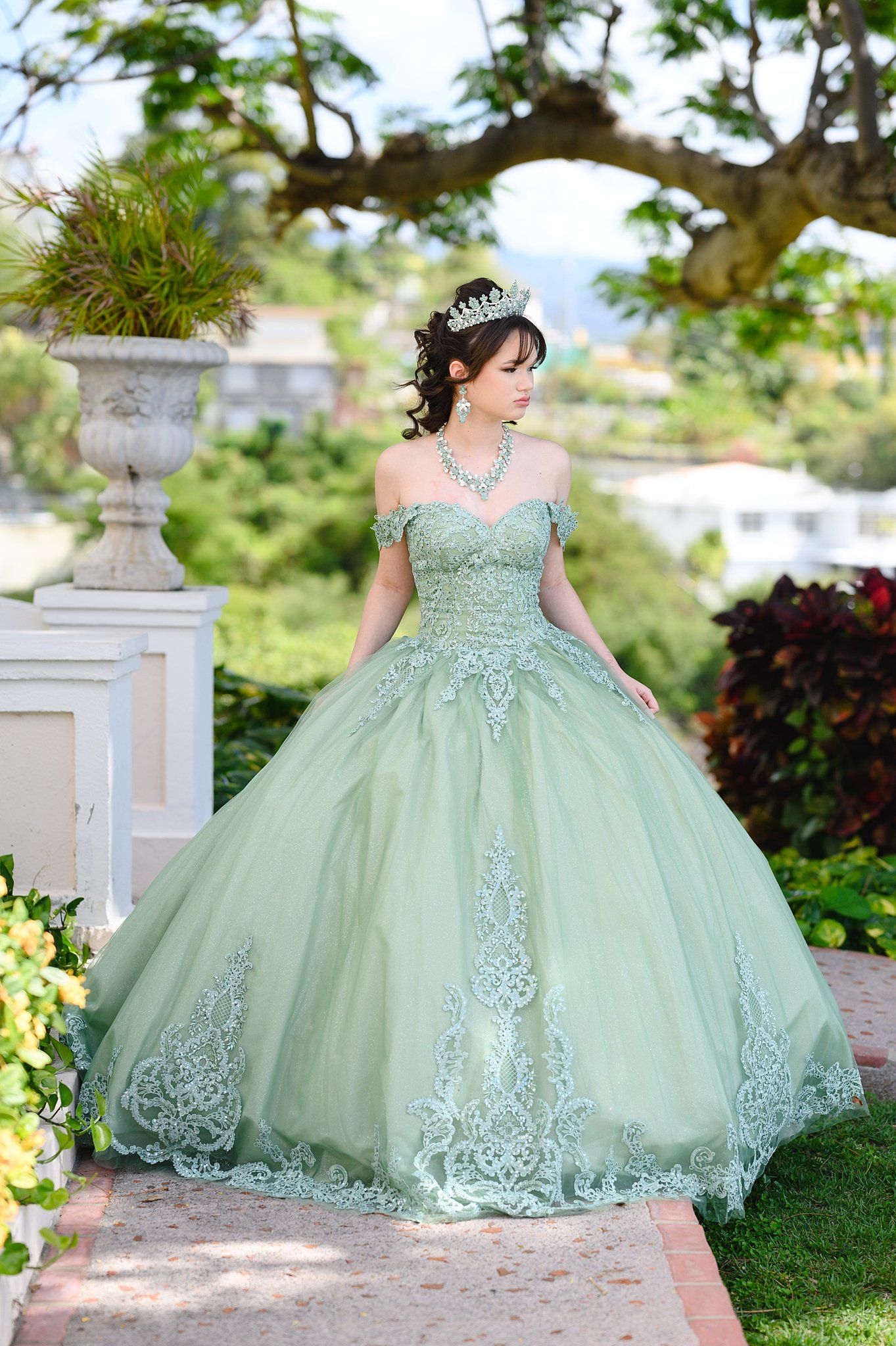Young woman in sage green off-shoulder quinceañera ball gown with lace appliqués and tiara poses outdoors.