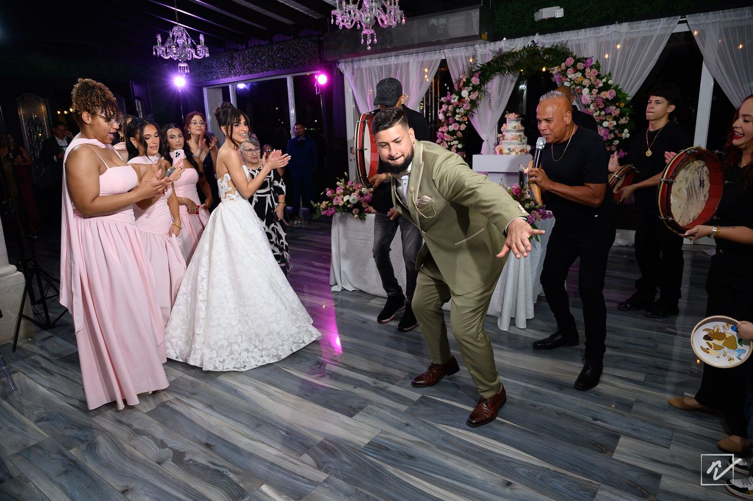 Groom in an olive green suit dances playfully while the bride in a white gown and bridesmaids in pink clap at a wedding reception.