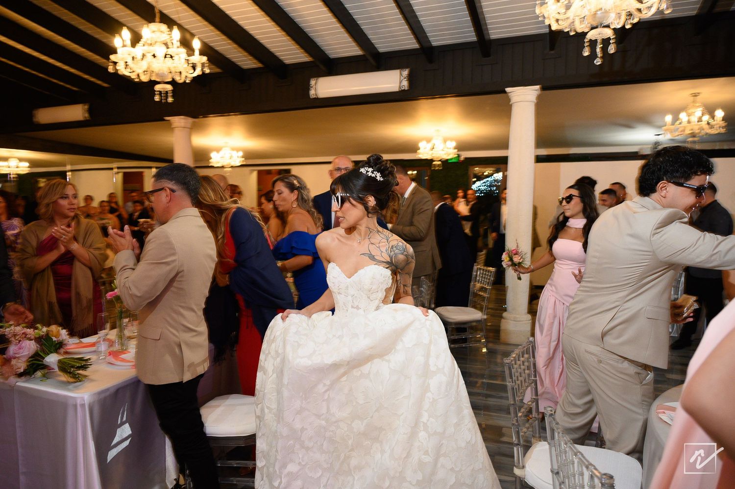 Bride in a white gown walks through a crowd of guests mingling at an elegant wedding reception under glowing chandeliers.