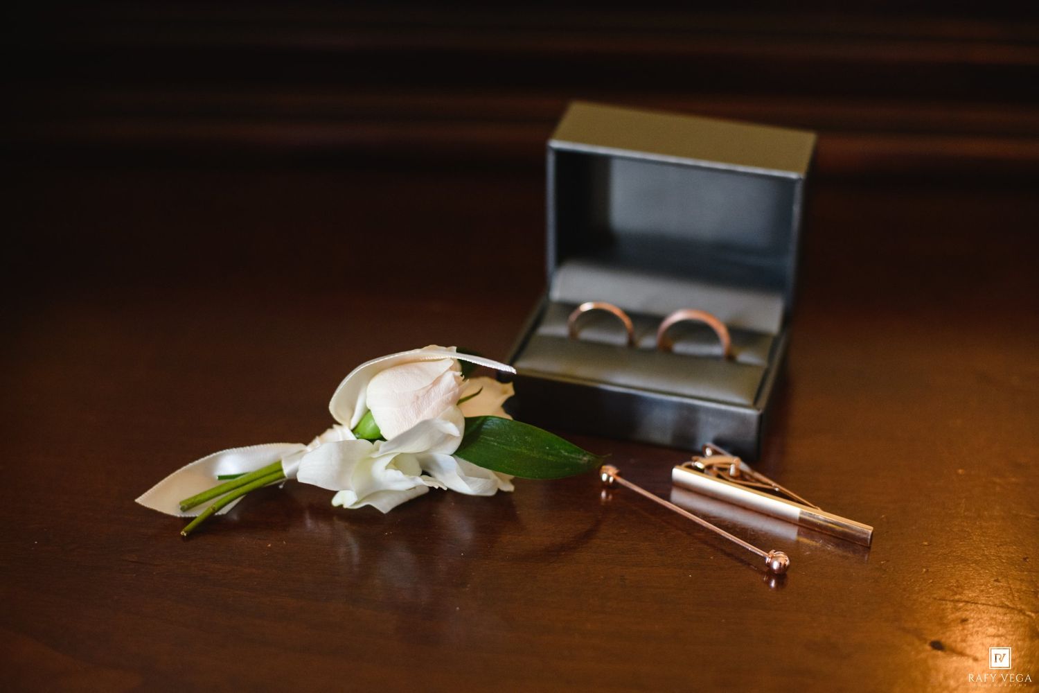 Wedding rings in a gray box beside a white rose boutonniere and gold tie clip on a wooden surface.