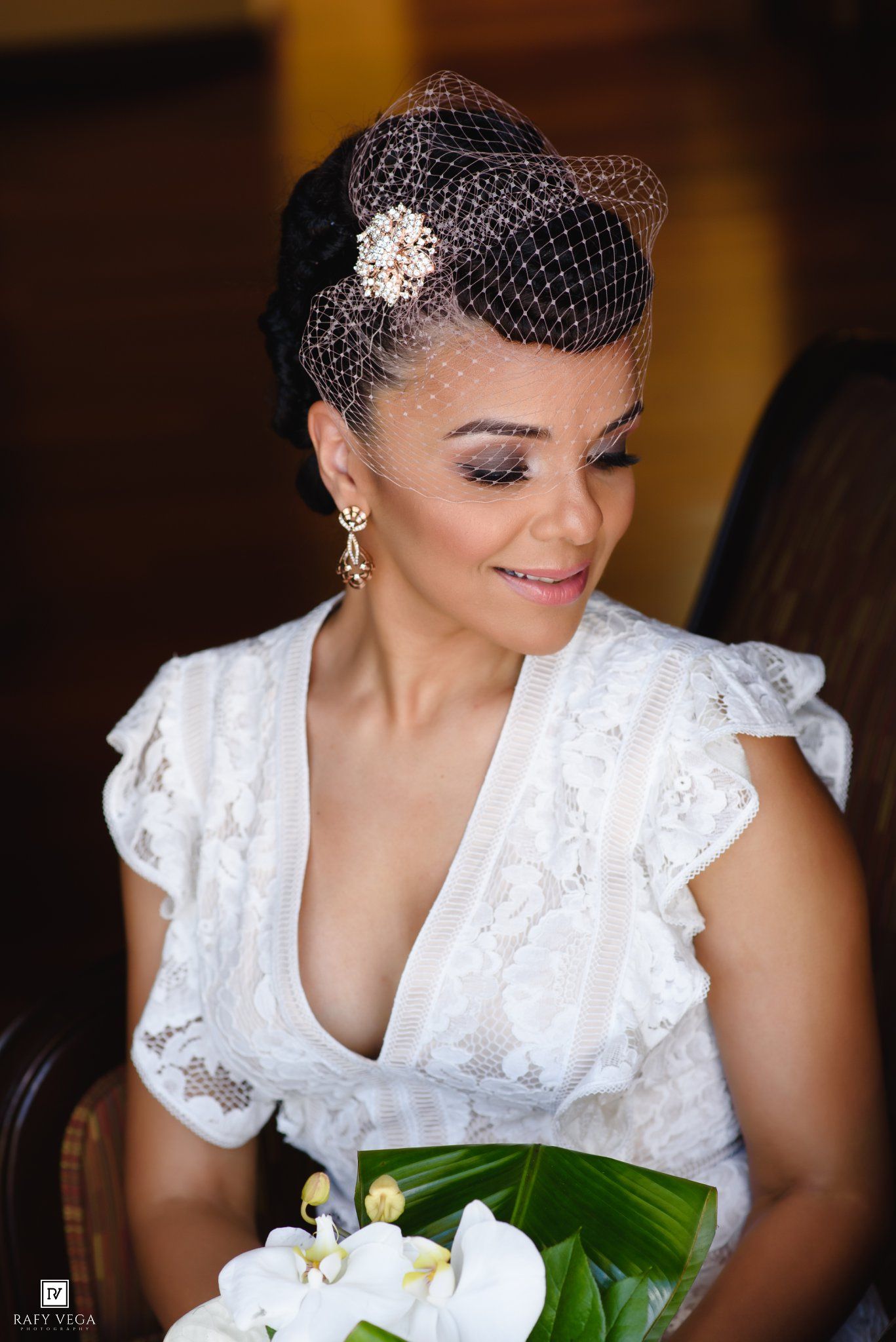 Elegant bride in white lace dress with birdcage veil, brooch hair accessory, and white orchid bouquet on her wedding day.