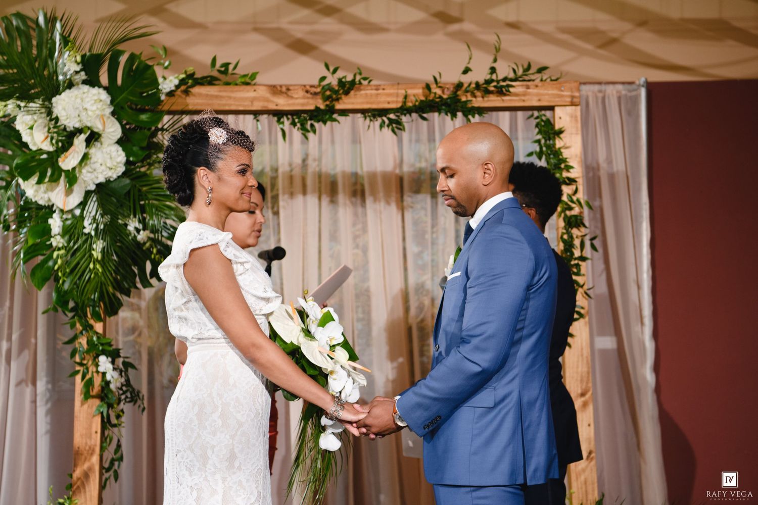 Bride and groom holding hands at wedding altar with white floral arch decor during outdoor ceremony.