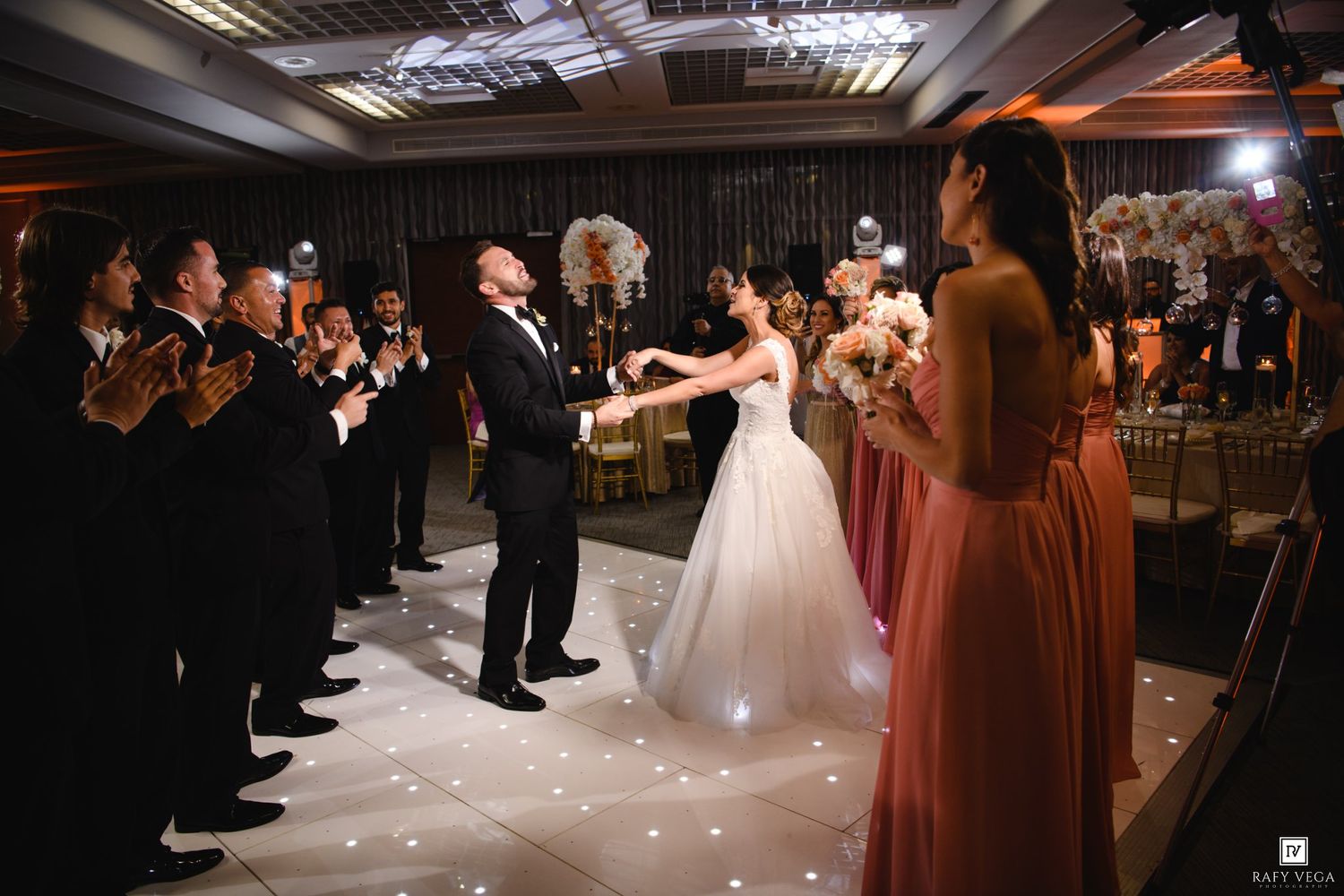 Bride and groom share first dance at elegant wedding reception while guests applaud and bridesmaids in rose gowns look on.