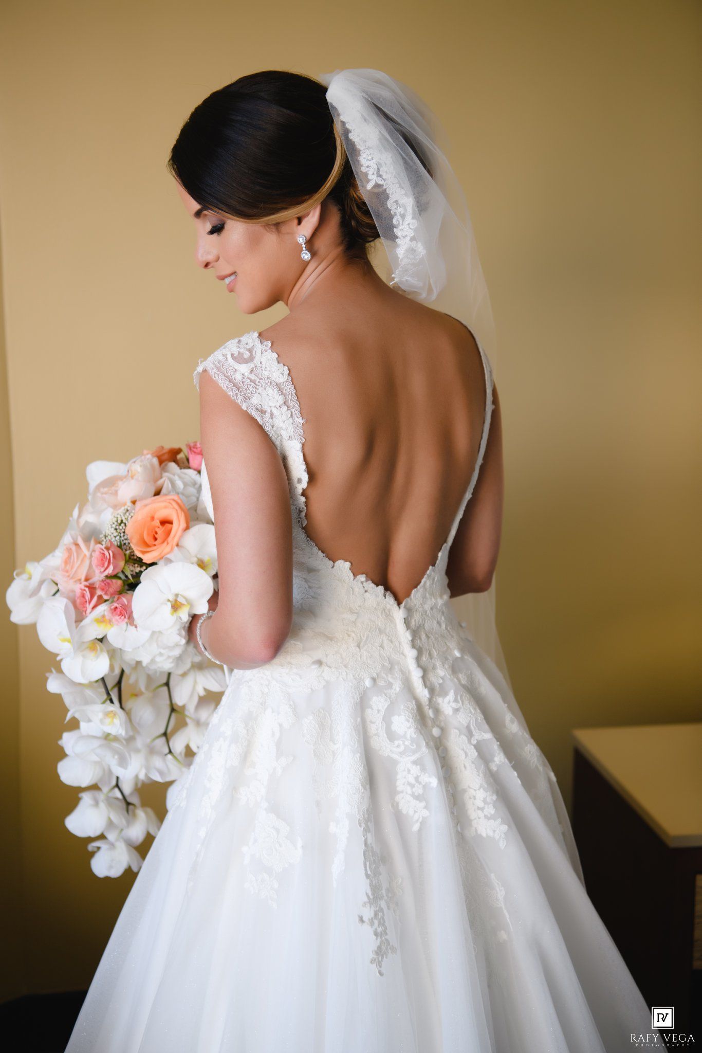 Bride in backless lace wedding gown holding cascading white orchid and peach rose bouquet, veil draped over updo hairstyle.