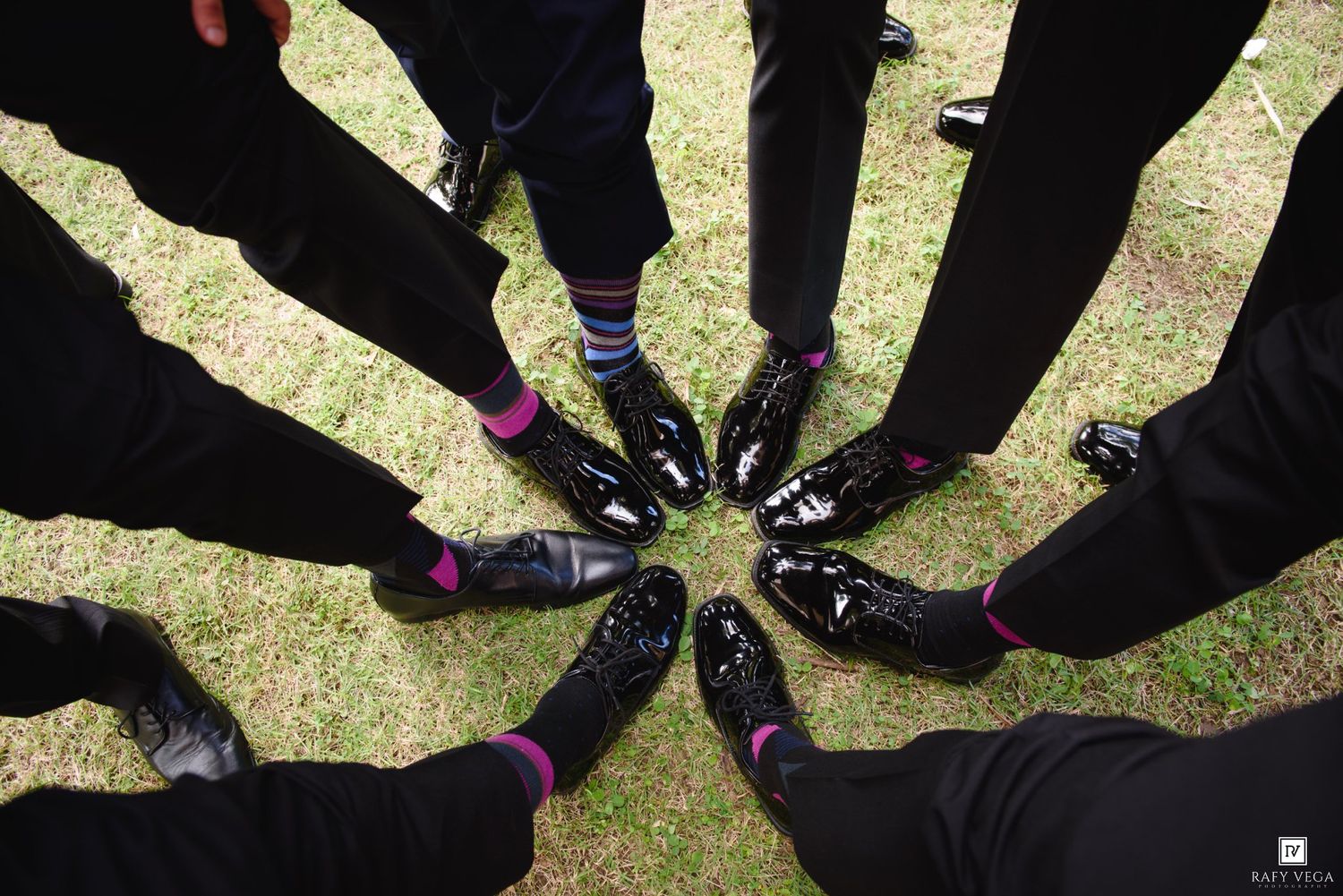 Groomsmen in black suits show off colorful socks and shiny dress shoes in a circle on green grass.