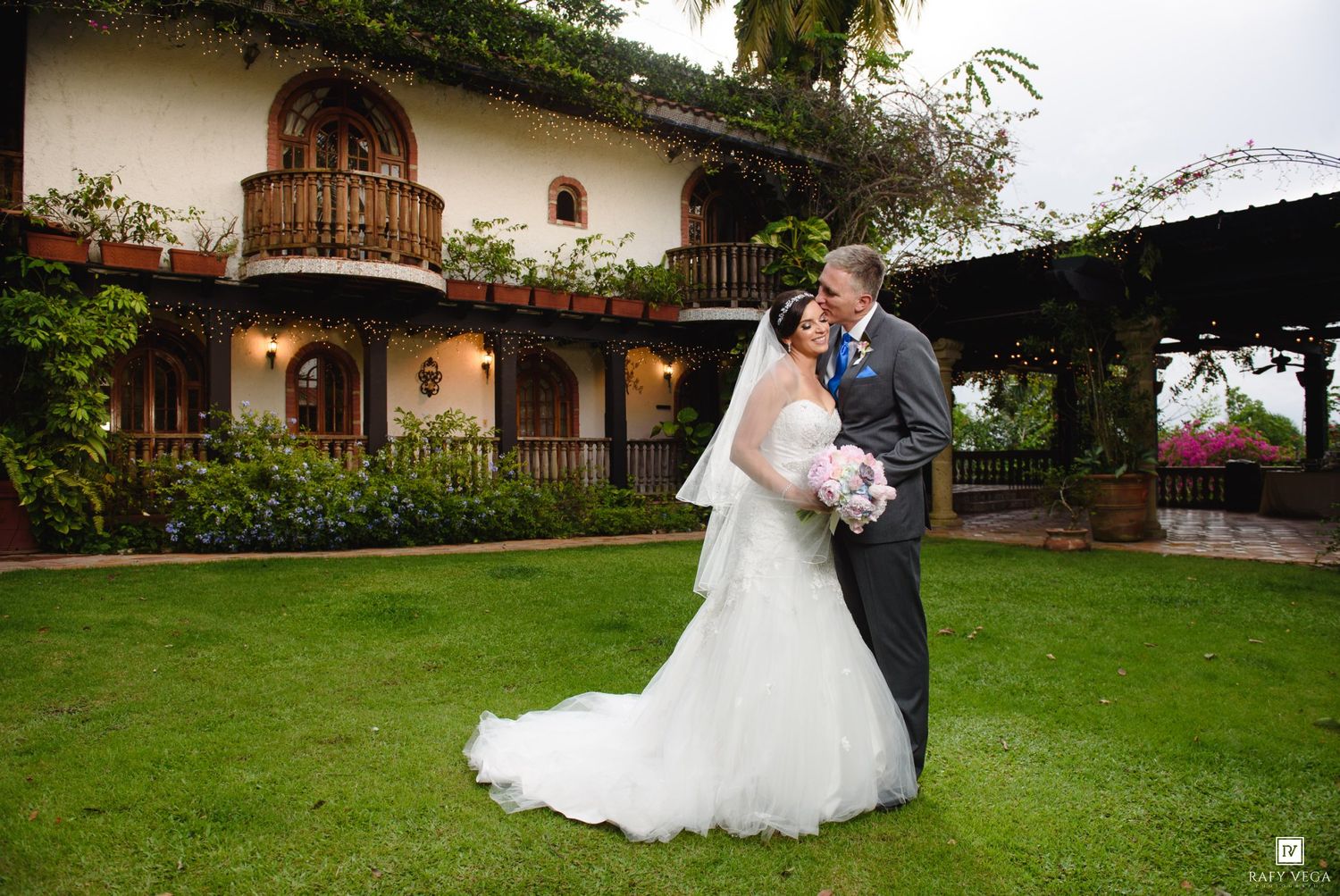 Bride and groom share a romantic moment on lush green lawn outside elegant Spanish-style wedding venue.