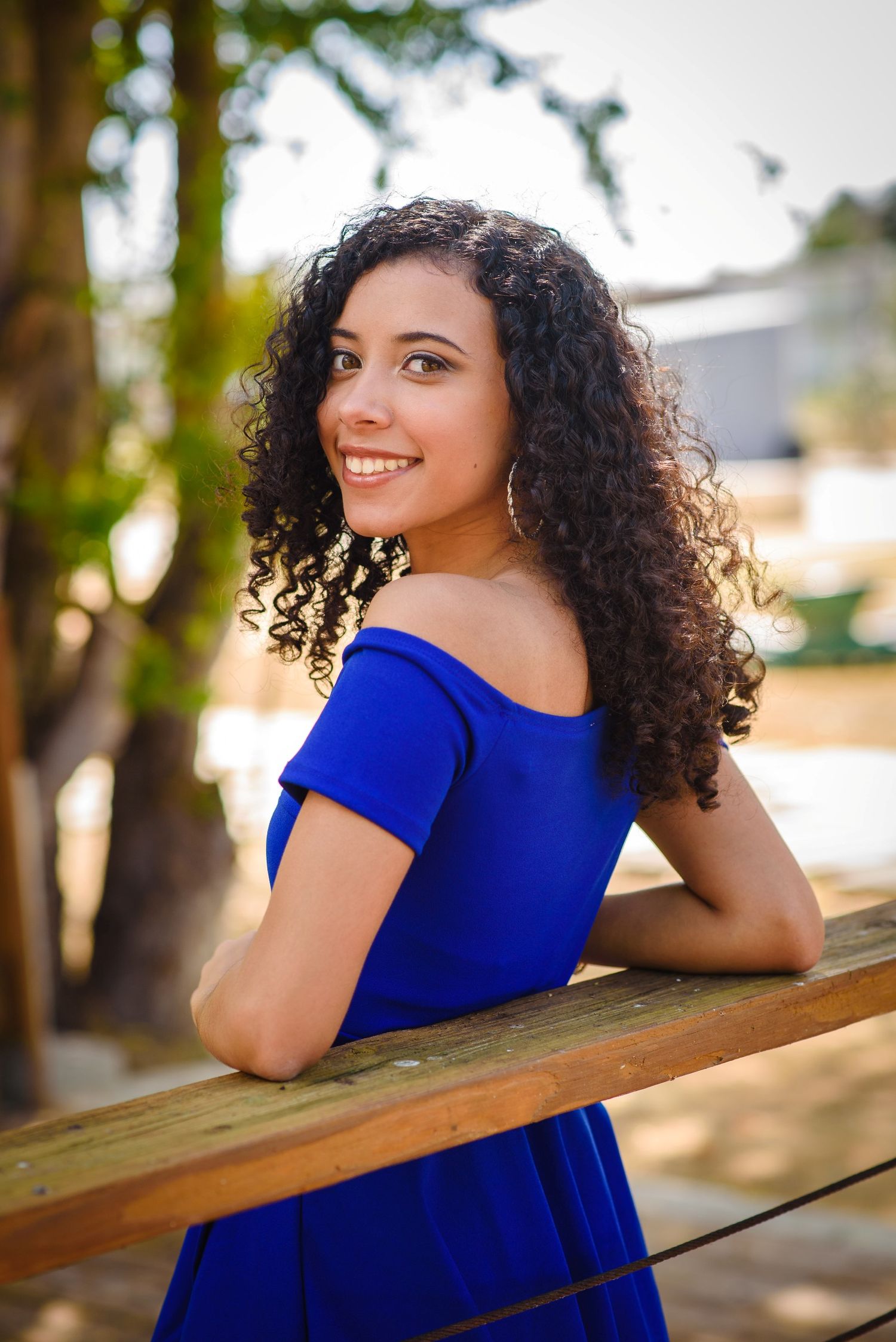 Young woman with curly hair smiling in a blue off-shoulder dress, leaning on a wooden railing outdoors.