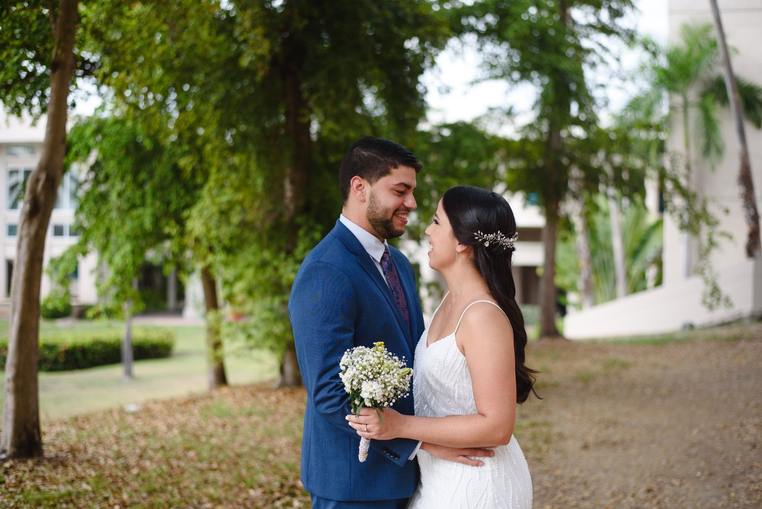 A bride and groom share a romantic moment outdoors, surrounded by lush trees, as she holds a white bouquet.
