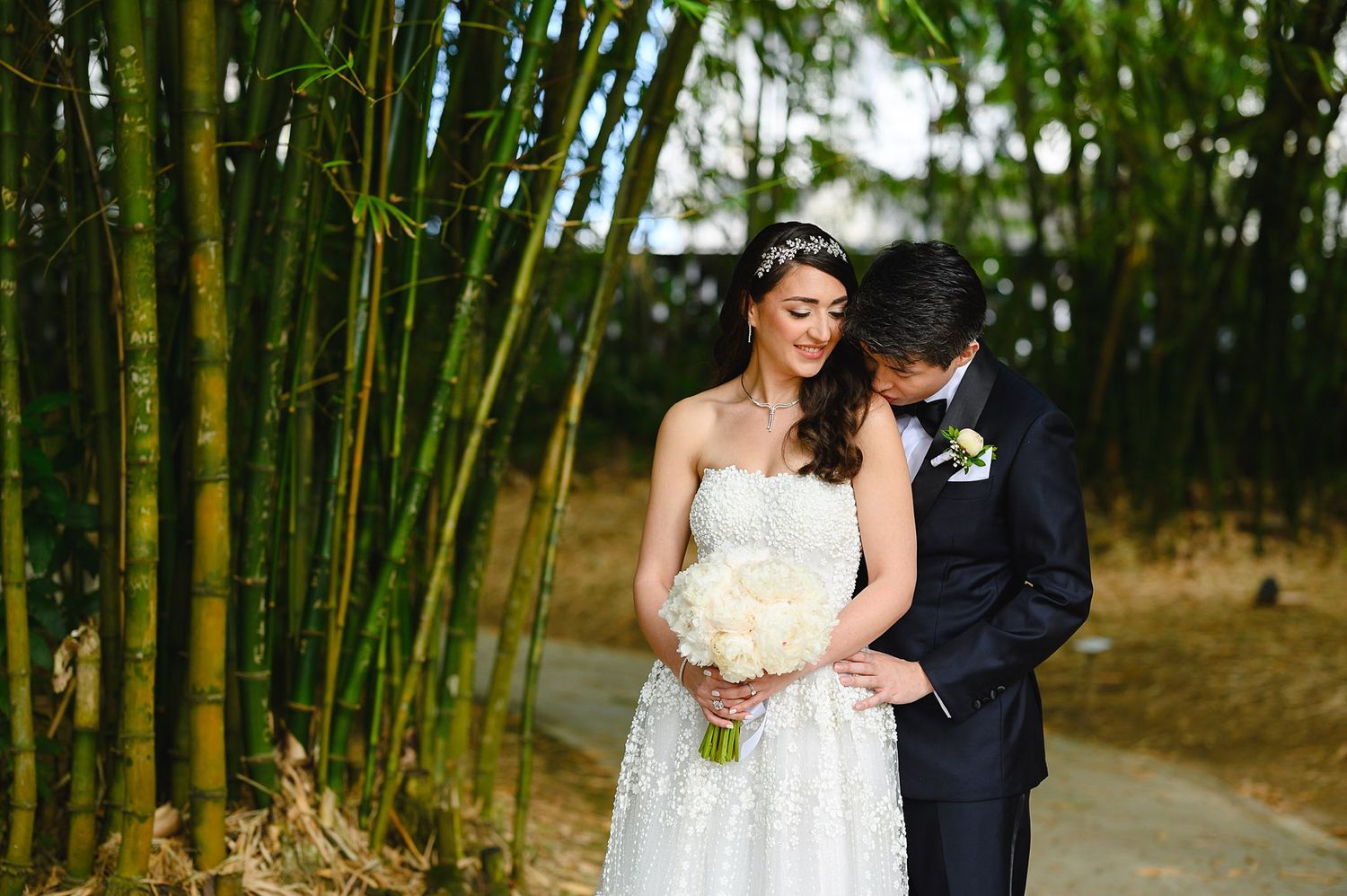 Bride and groom share intimate moment among bamboo trees, bride holding white bouquet in lace wedding gown.