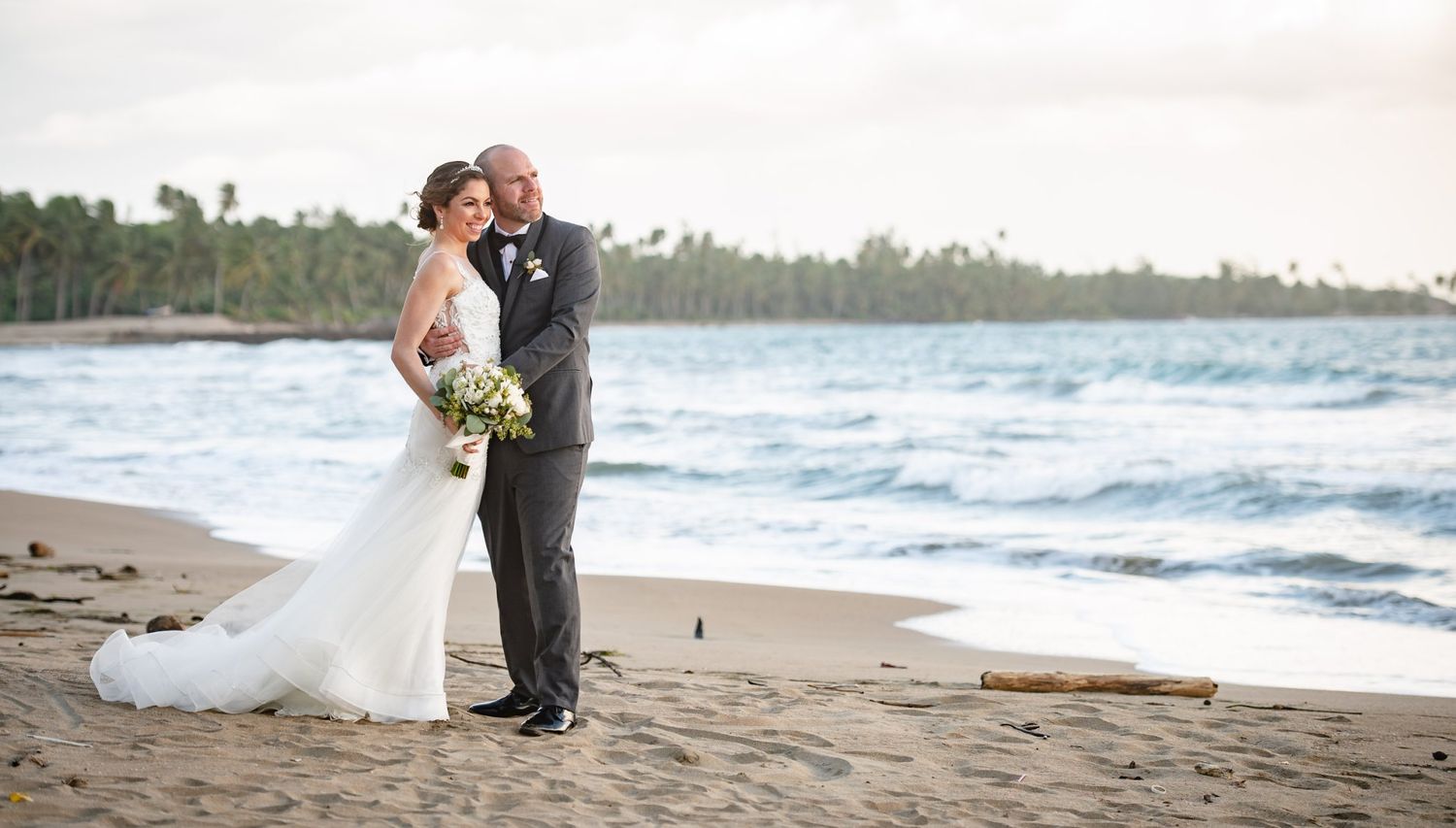 Bride and groom share a romantic kiss on a tropical beach at sunset, with palm trees and ocean waves in the background.