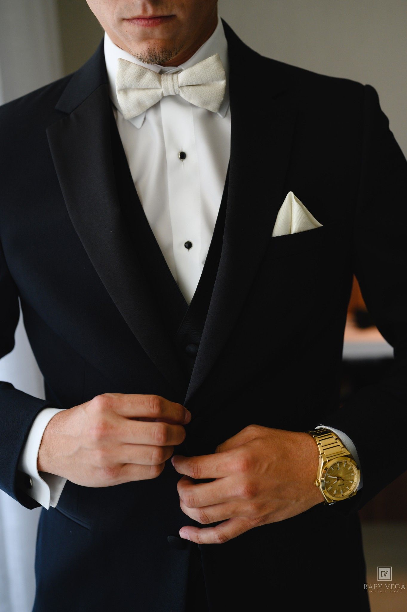 Groom in black tuxedo with white bow tie and gold watch buttoning jacket before wedding ceremony.