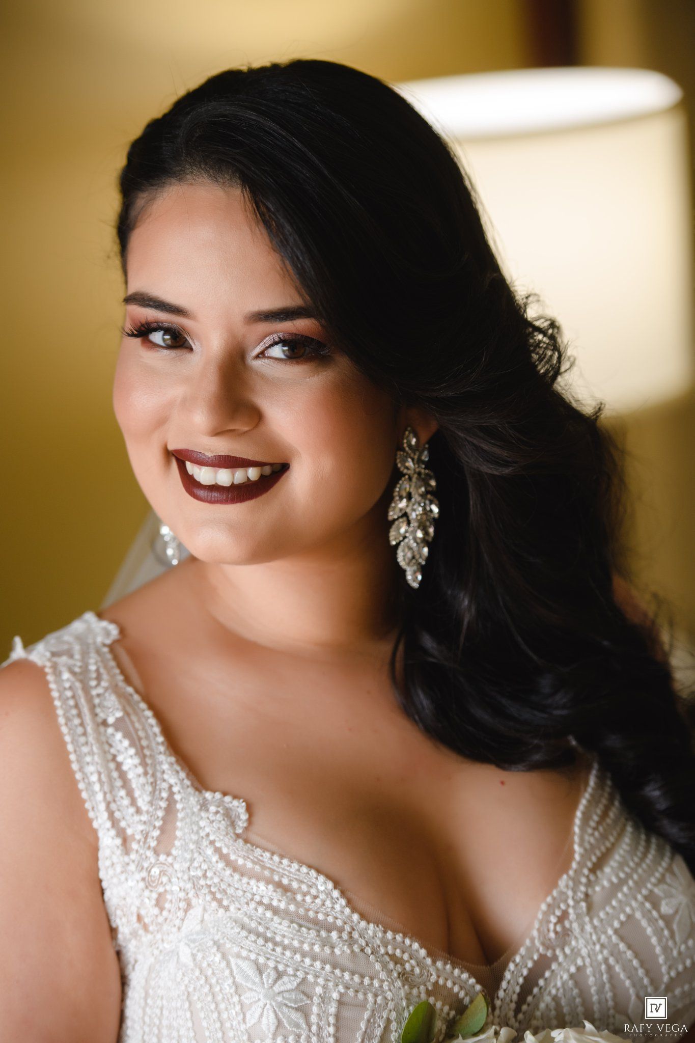 Radiant bride in pearl-beaded gown with chandelier earrings, dark lips, and side-swept hair, smiling beautifully.