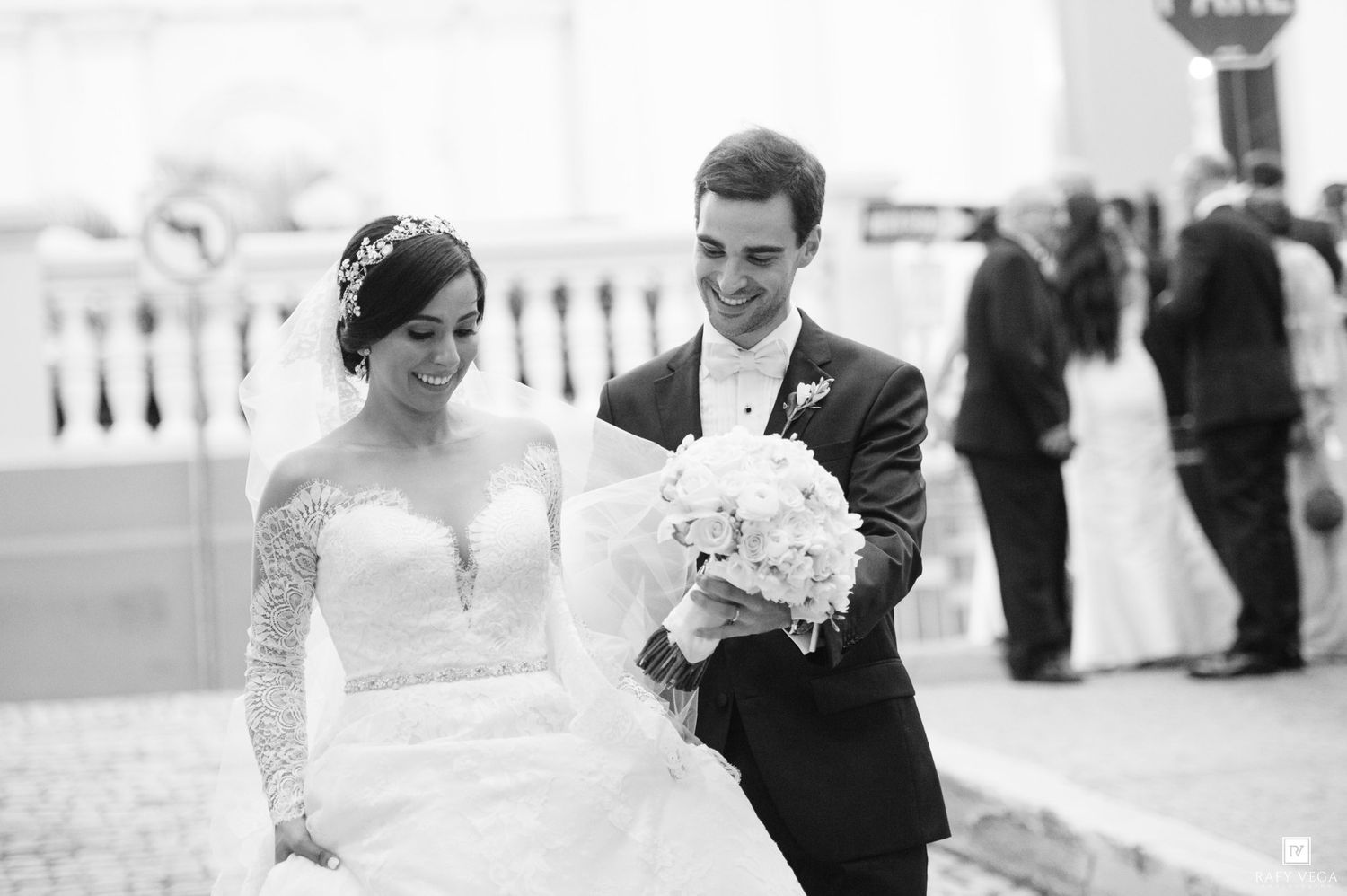 Black and white wedding photo of smiling bride in lace gown and groom in suit walking together outdoors.
