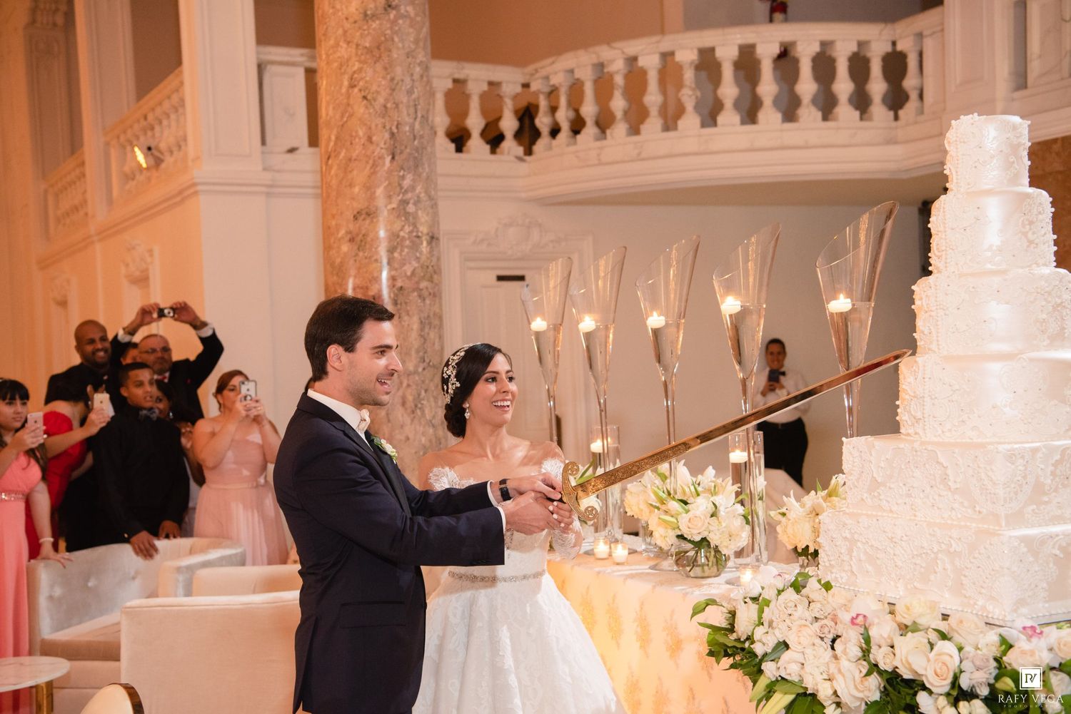 Bride and groom cutting elegant white tiered wedding cake at luxurious ballroom reception with guests celebrating.