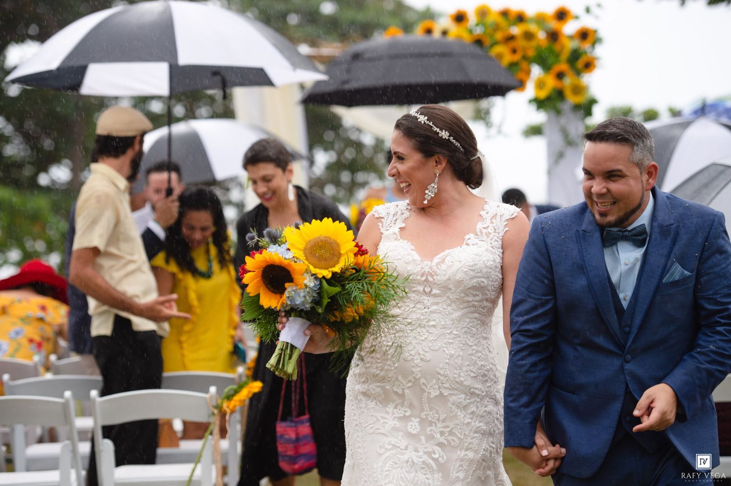 Bride holding sunflower bouquet and groom in blue suit walk down rainy outdoor wedding aisle with guests holding umbrellas.