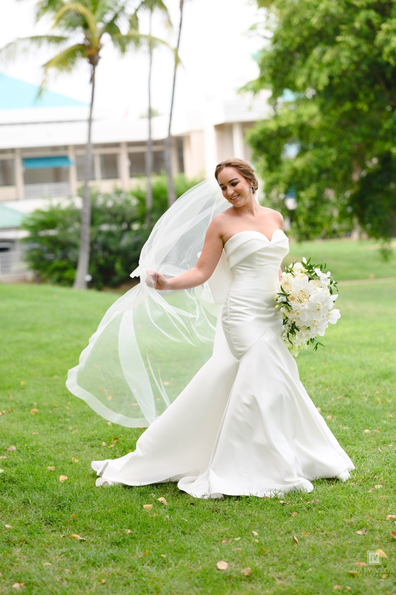 Bride in white mermaid gown twirling veil on green lawn, holding white floral bouquet, with tropical trees behind.