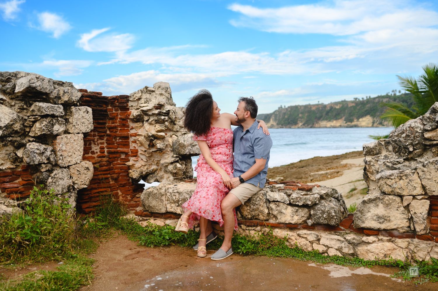 Couple sharing a romantic moment by ancient stone ruins near a tropical beach under a blue sky.