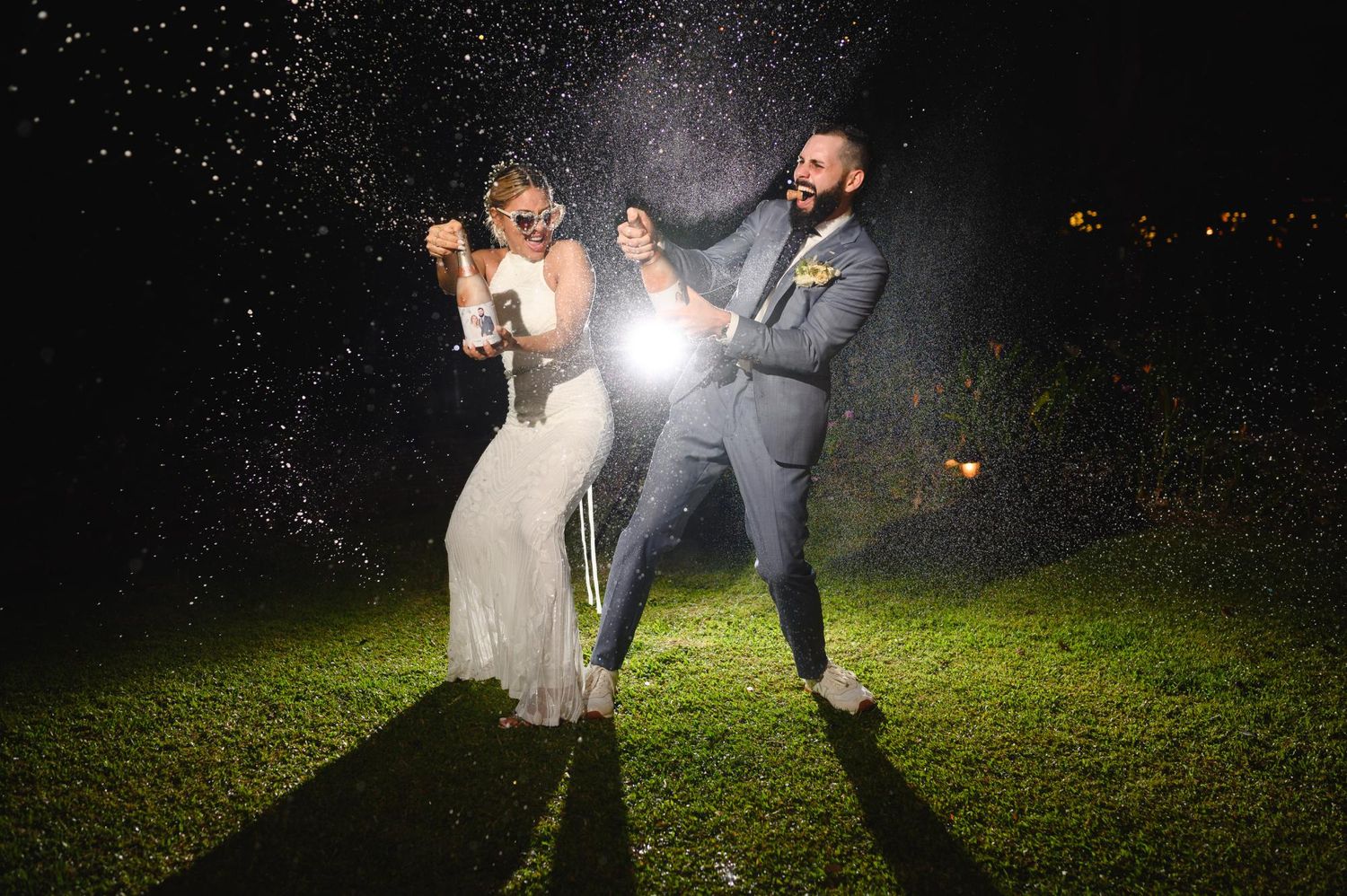 Bride and groom celebrating at night, spraying champagne outdoors on green grass in wedding attire.