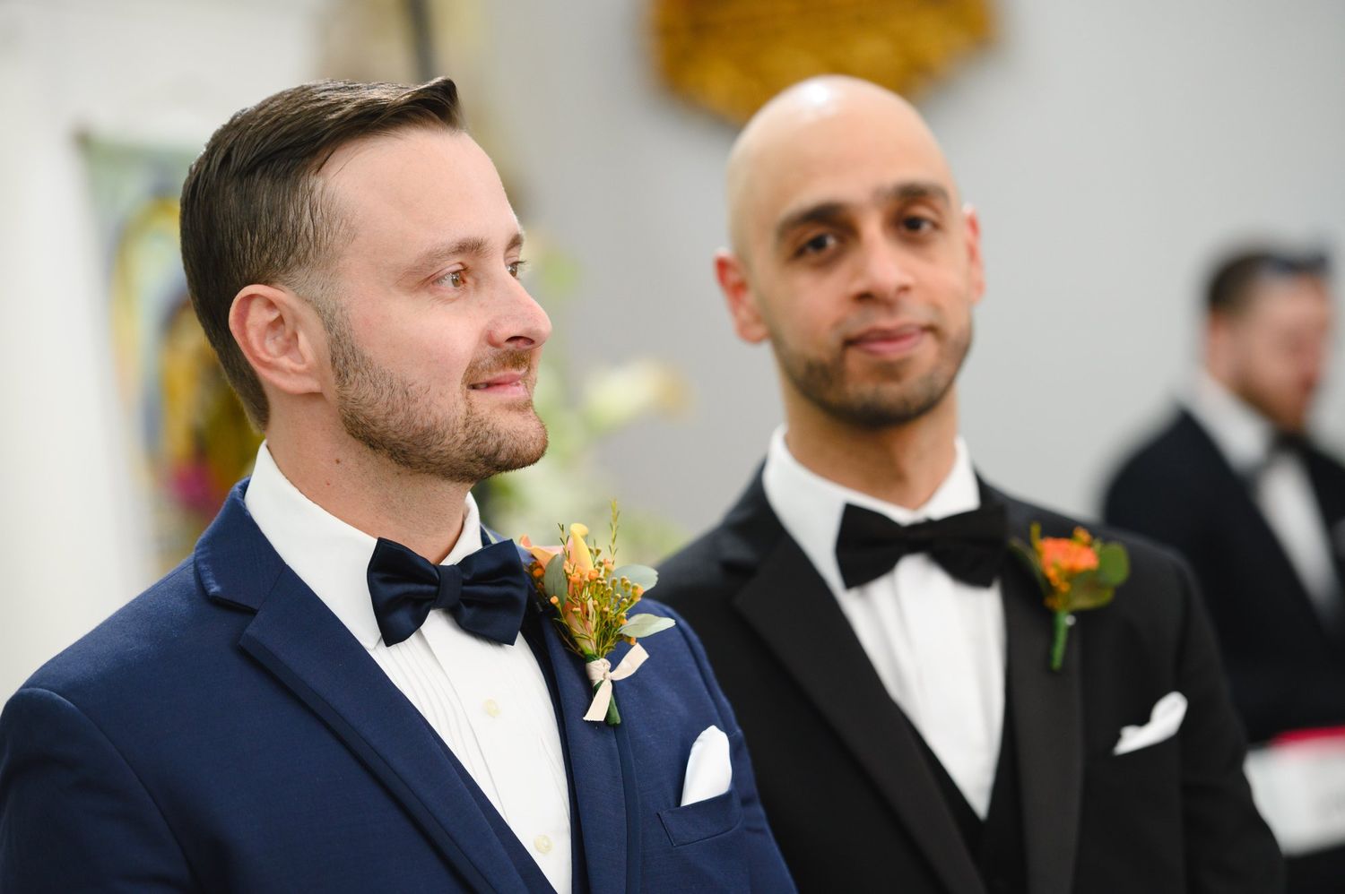Two grooms in tuxedos with boutonnieres stand at the altar during a wedding ceremony.